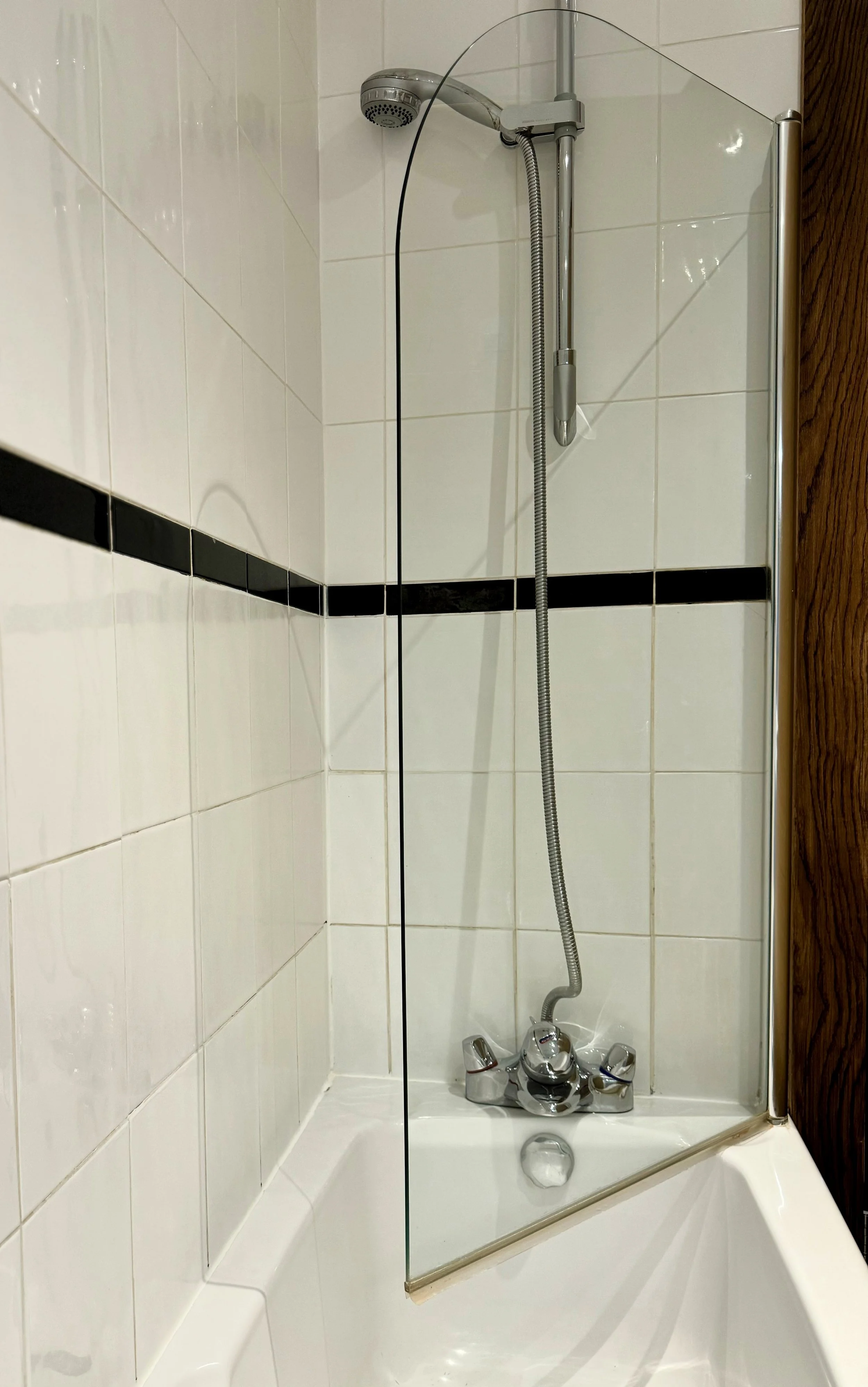 A picture of a shower area with white tiled walls, a black tiled accent strip, a chrome showerhead, a flexible hose, a glass shower wall, and a brown wooden panel to the right side.
