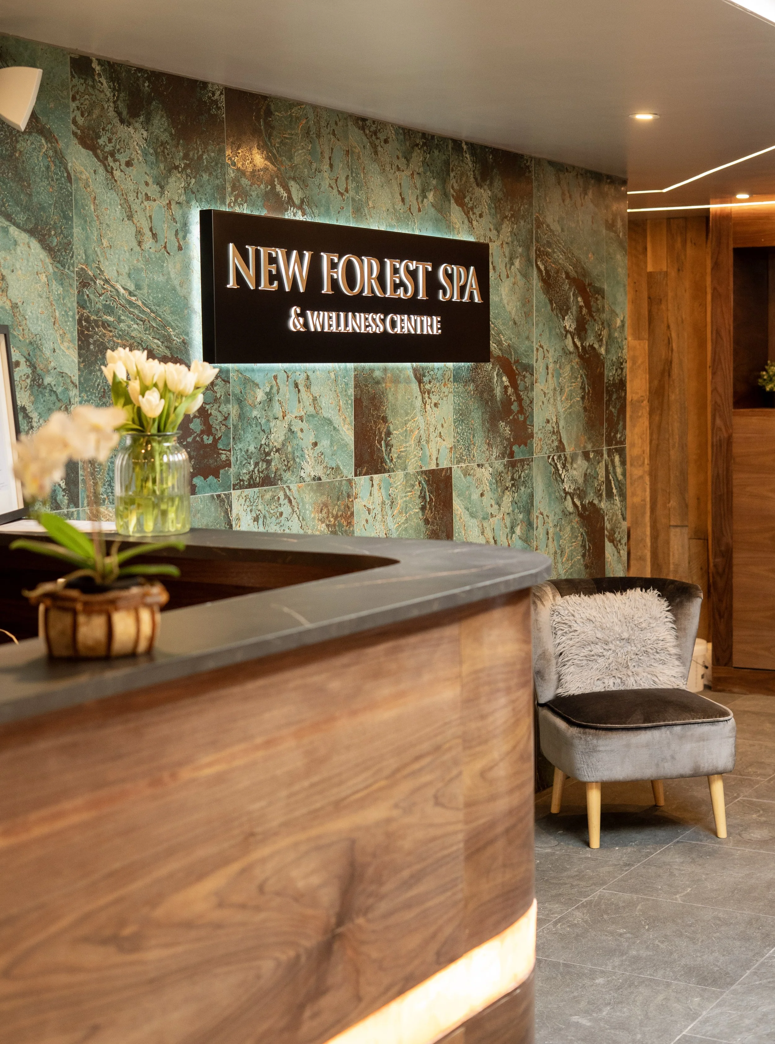 Reception area of New Forest Spa & Wellness Centre with a marble wall, wooden counter, and a gray chair with a fluffy cushion.