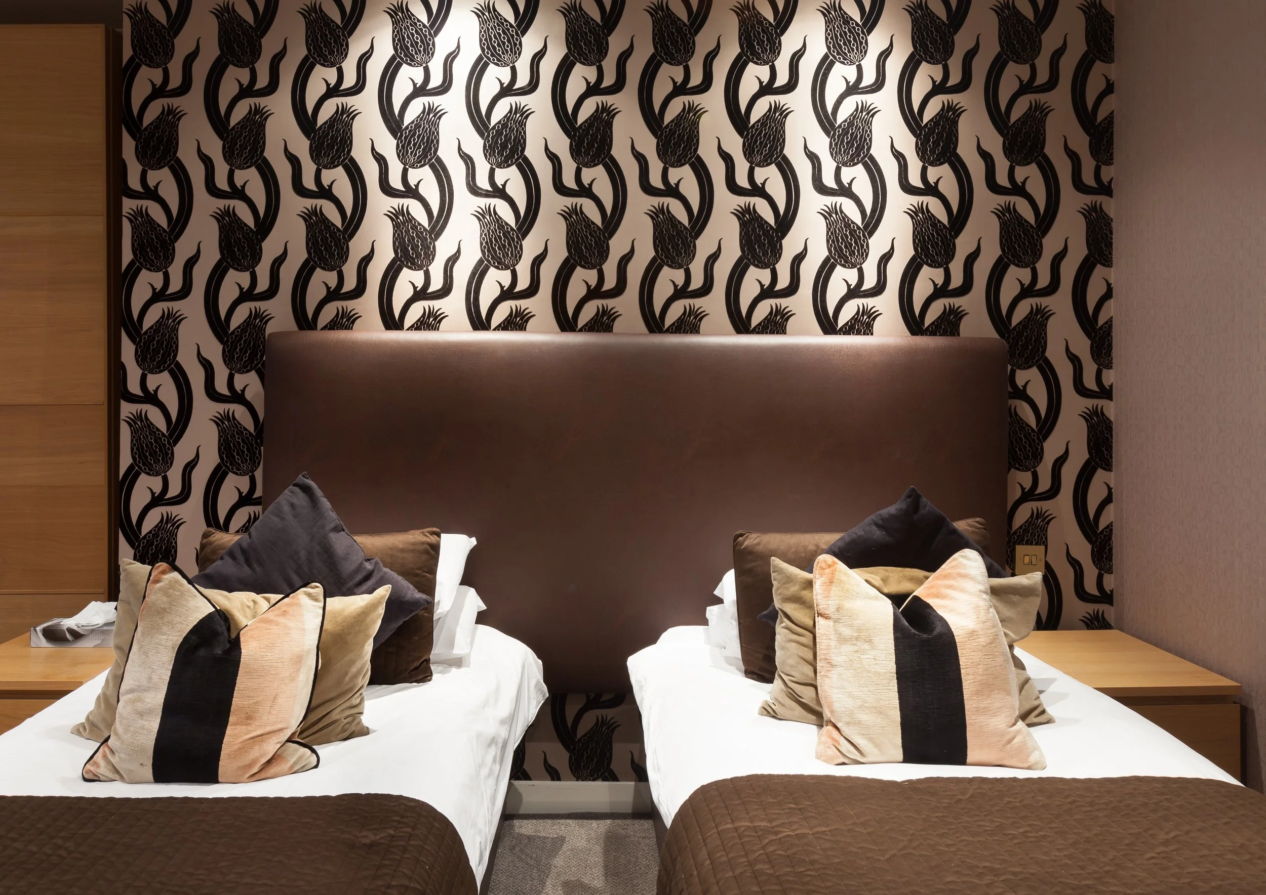Two single beds in a hotel room with brown headboards, decorated with multiple pillows in neutral colors, and a wallpaper featuring black stylized floral patterns.
