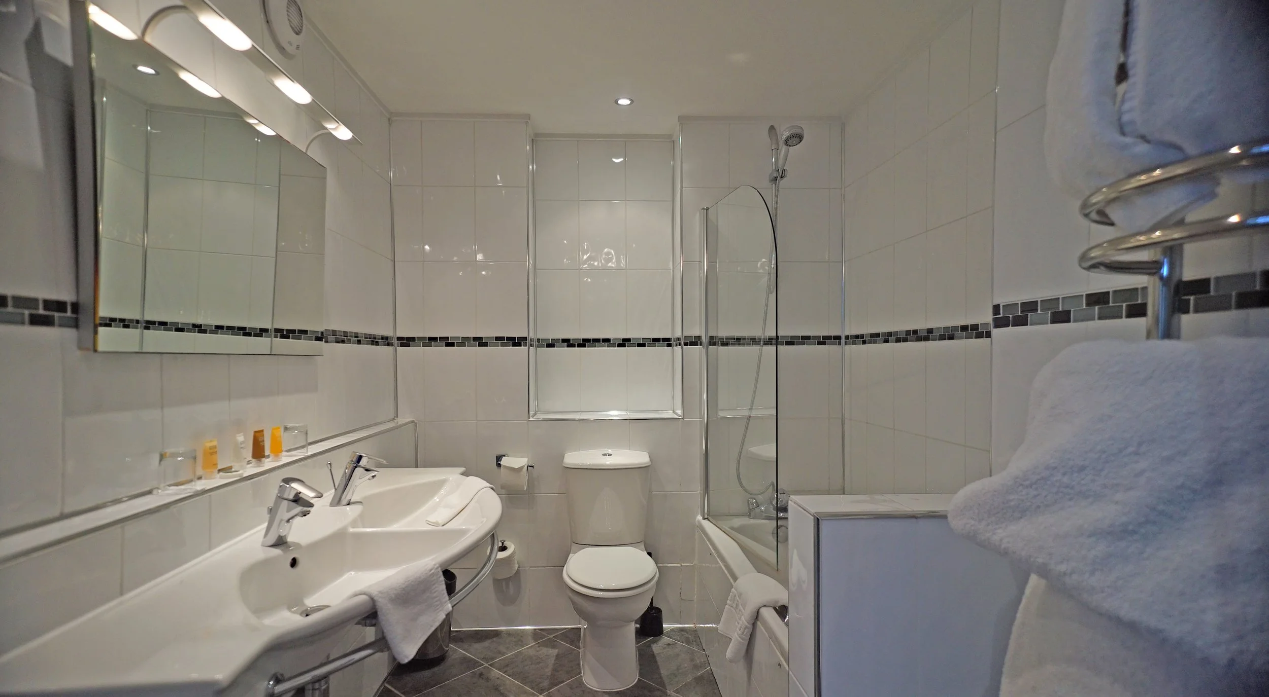 Bathroom with white tiled walls, a large mirror with lights, dual sinks with towels, a toilet, a bathtub with a glass shower screen, and a heated towel rack with towels.