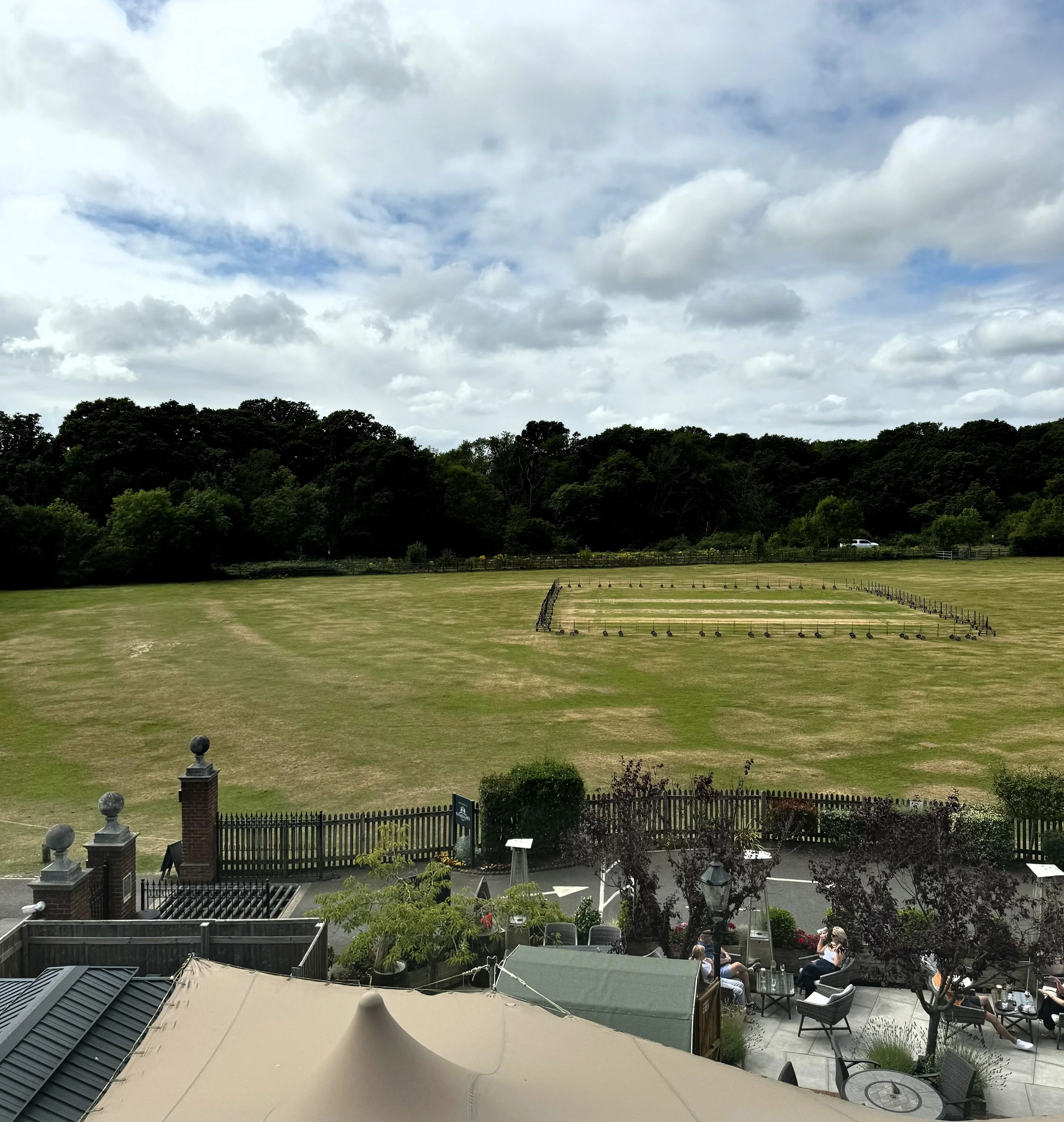 A scenic outdoor view with a large grassy field, a fence, and a group of people sitting at tables under umbrellas in the foreground. The background features a tree line under a partly cloudy sky.