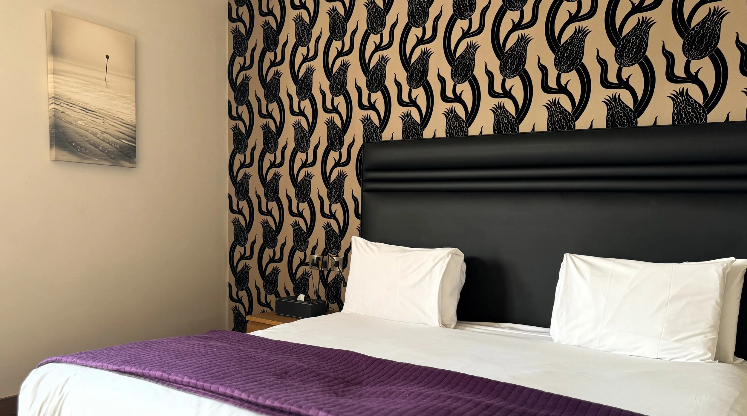 A hotel room with a bed that has white sheets, two white pillows, a purple bed runner, and a black headboard. There is a patterned wallpaper with black abstract floral designs on beige background behind the bed. A nightstand with a tissue box and a b