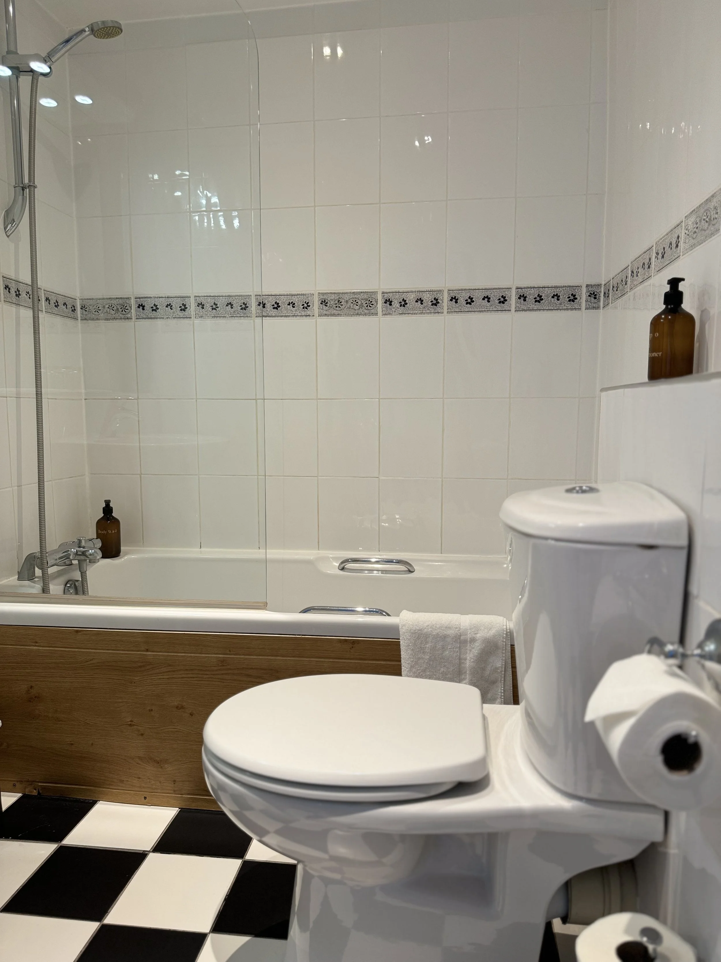 A bathroom with a toilet, a bathtub with a shower, and a black and white checkered floor. The bathtub has a glass partition and contains brown bottles, likely for soap or shampoo, placed on the edge.