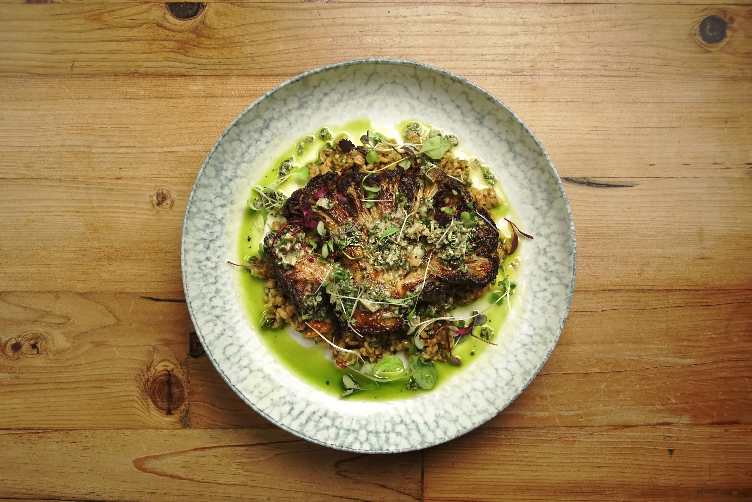 A dish featuring a roasted piece of meat, possibly chicken, served over a bed of grains and topped with microgreens. The dish is drizzled with a vibrant green sauce and garnished with herbs, all on a textured, light-colored plate placed on a wooden surface.