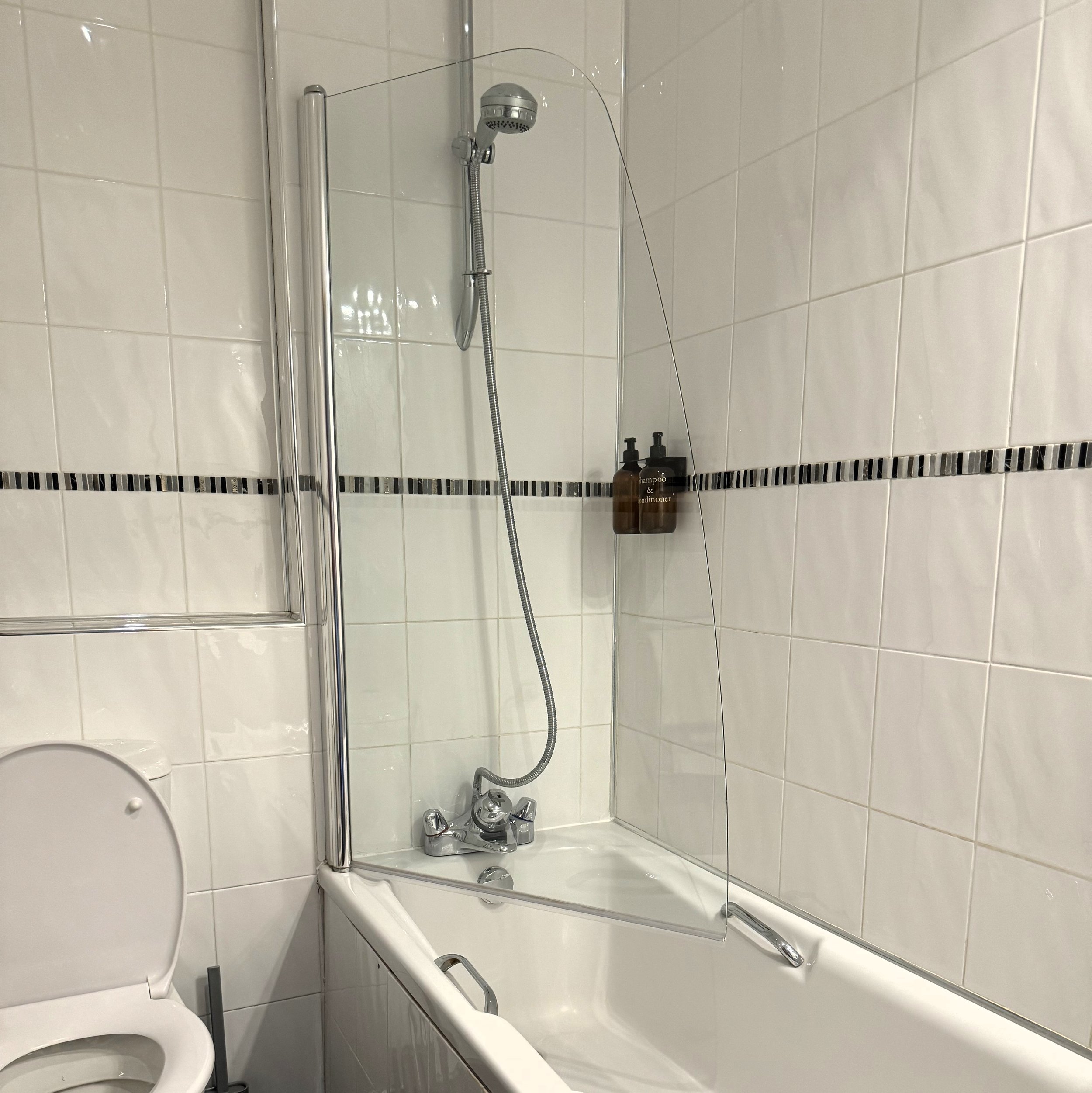 Bathroom with white tiled walls, a bathtub with a glass shower screen, a wall-mounted shower head, and two brown soap dispensers labeled 'Shampoo & Conditioner' in the corner.