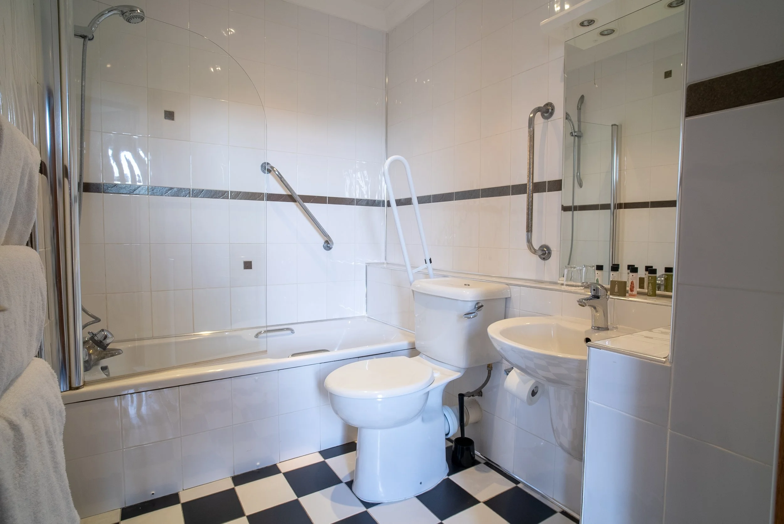 A compact bathroom with a bathtub, toilet, sink, and mirror, equipped with safety grab bars and a foldable bath chair.