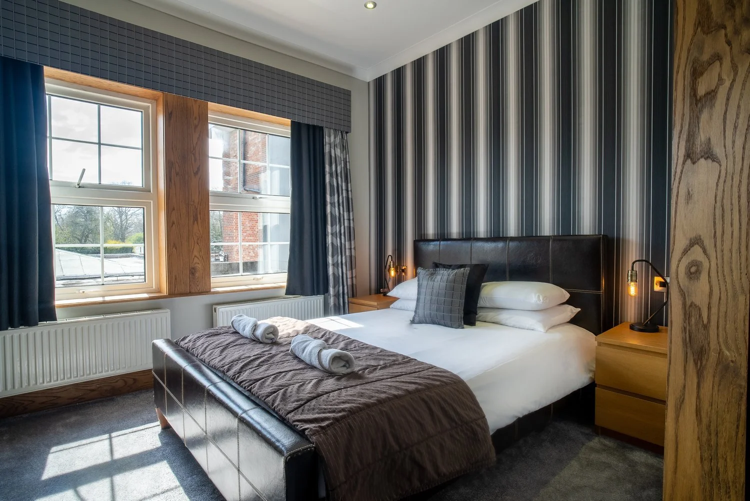 A bedroom with a large bed, striped wallpaper, and two windows with blue and patterned curtains. The bed has white linens, a plaid pillow, and rolled towels at the foot. Two nightstands with lamps are on each side of the bed.