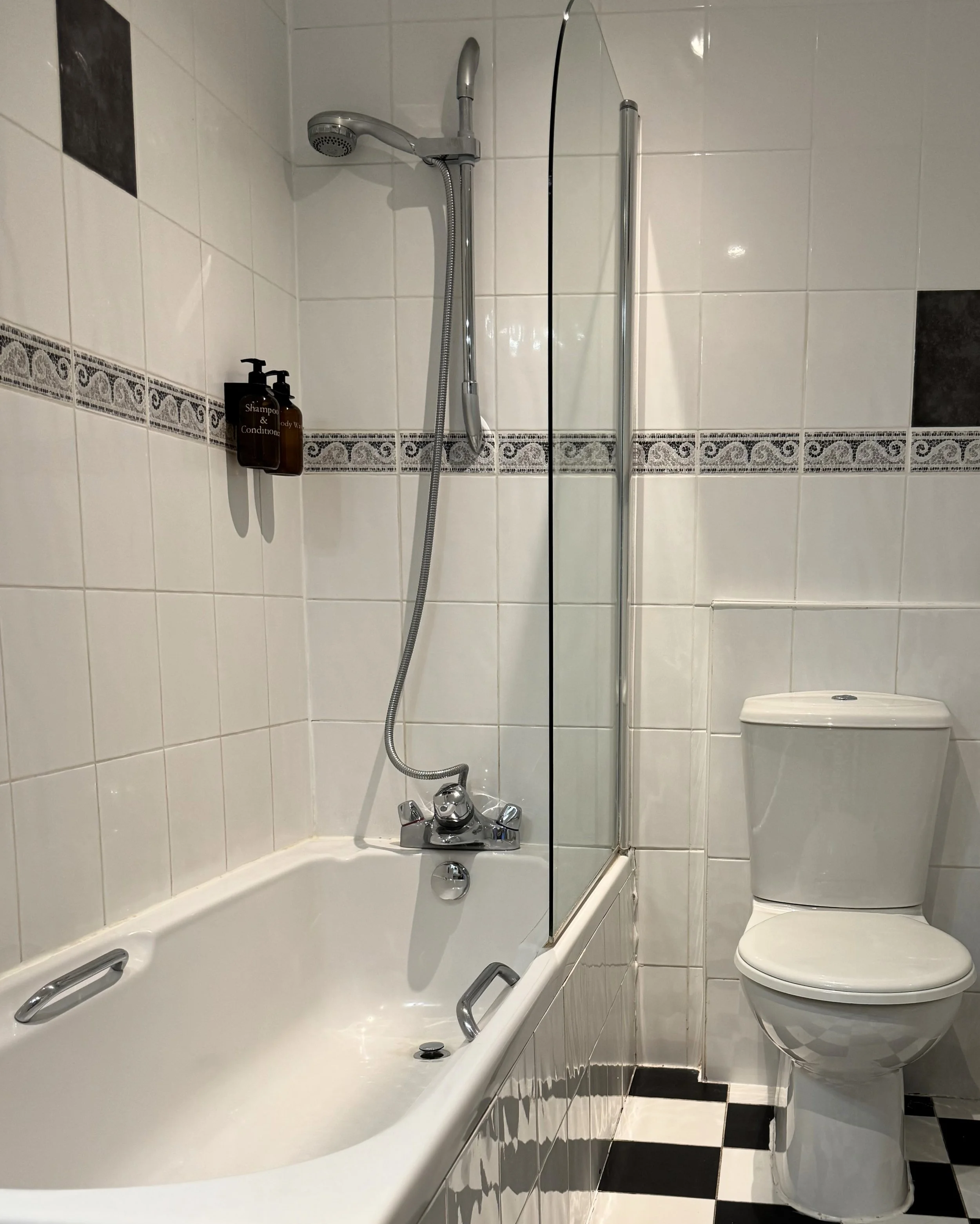 Bathroom with a bathtub, shower, and toilet, featuring white tiled walls with decorative black and grey border tiles, black and white checkered floor, and two brown soap dispensers on the wall above the bathtub.