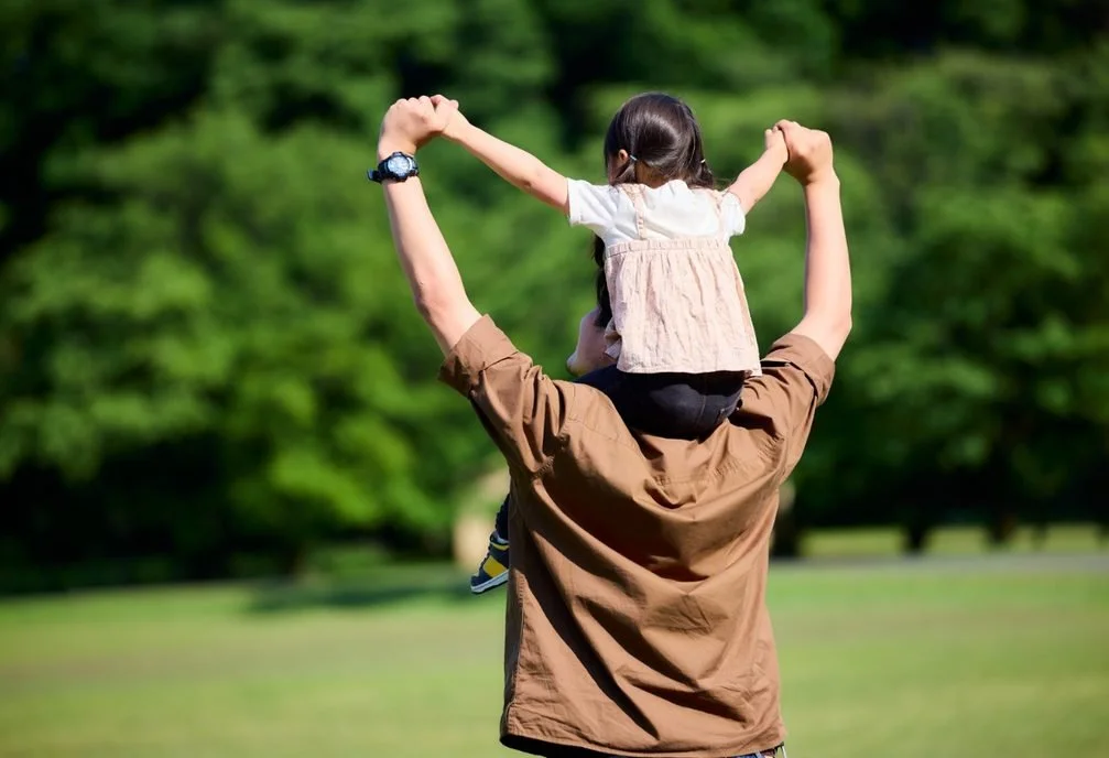 A man carrying a young girl on his shoulders in a park.