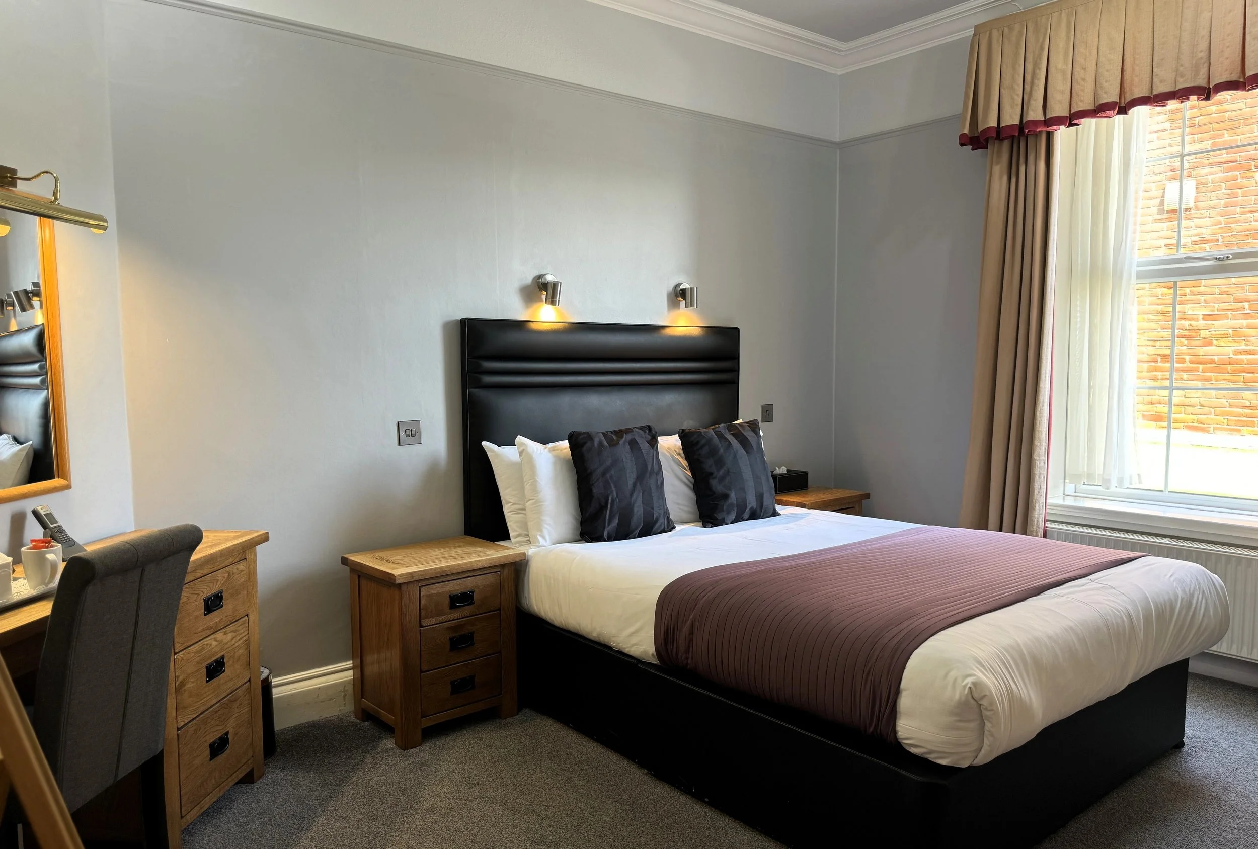 A tidy hotel bedroom with a king-sized bed, black headboard, white linens, and black pillows. Next to the bed are wooden nightstands, and a window with beige and red valance curtains reveals a brick building outside.