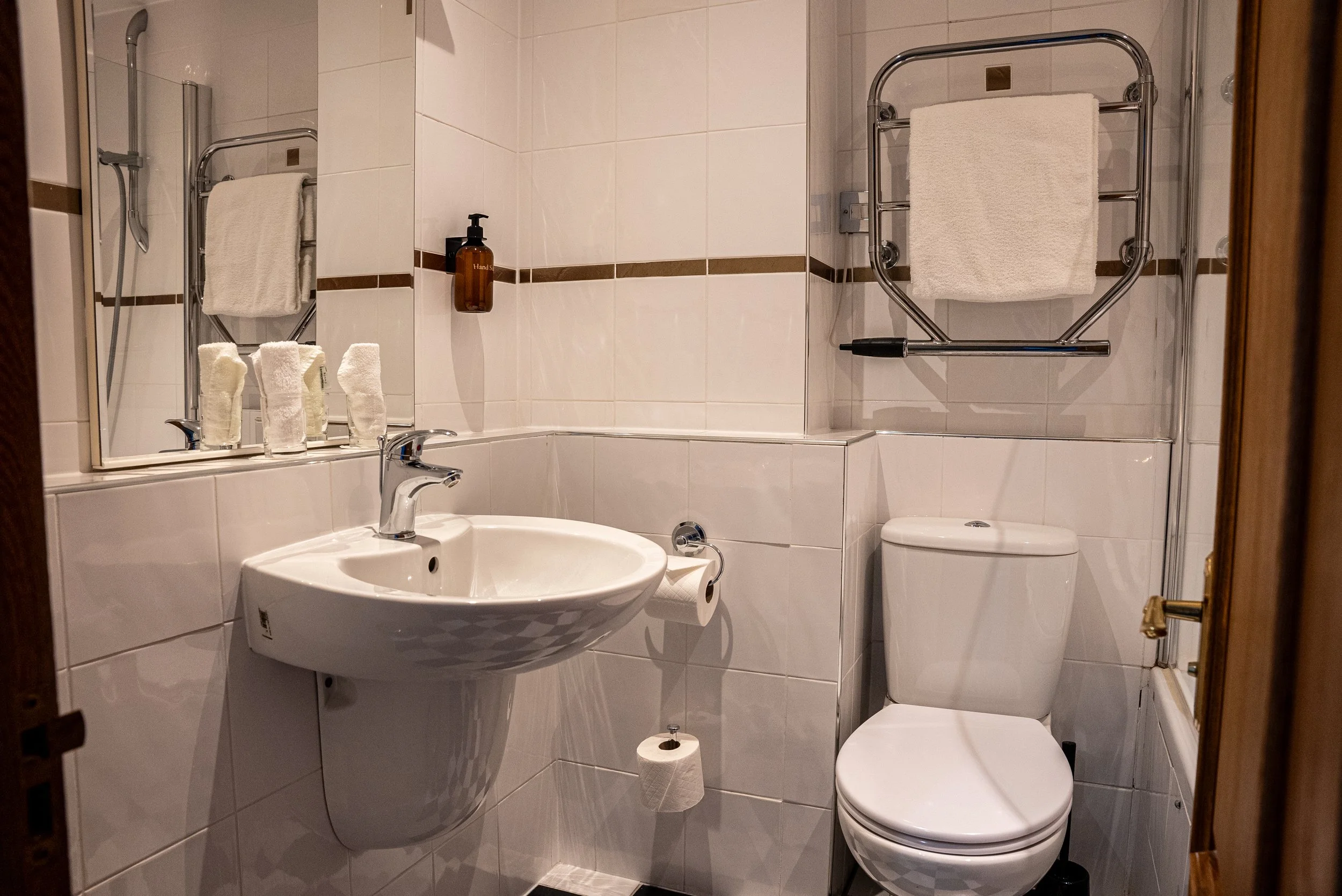 A small bathroom with white tiled walls, a mirror, a sink with a chrome faucet, a toilet, and a heated towel rack with a towel on it. There are three rolled towels and a soap dispenser on the mirror ledge, and toilet paper hanging on the wall.