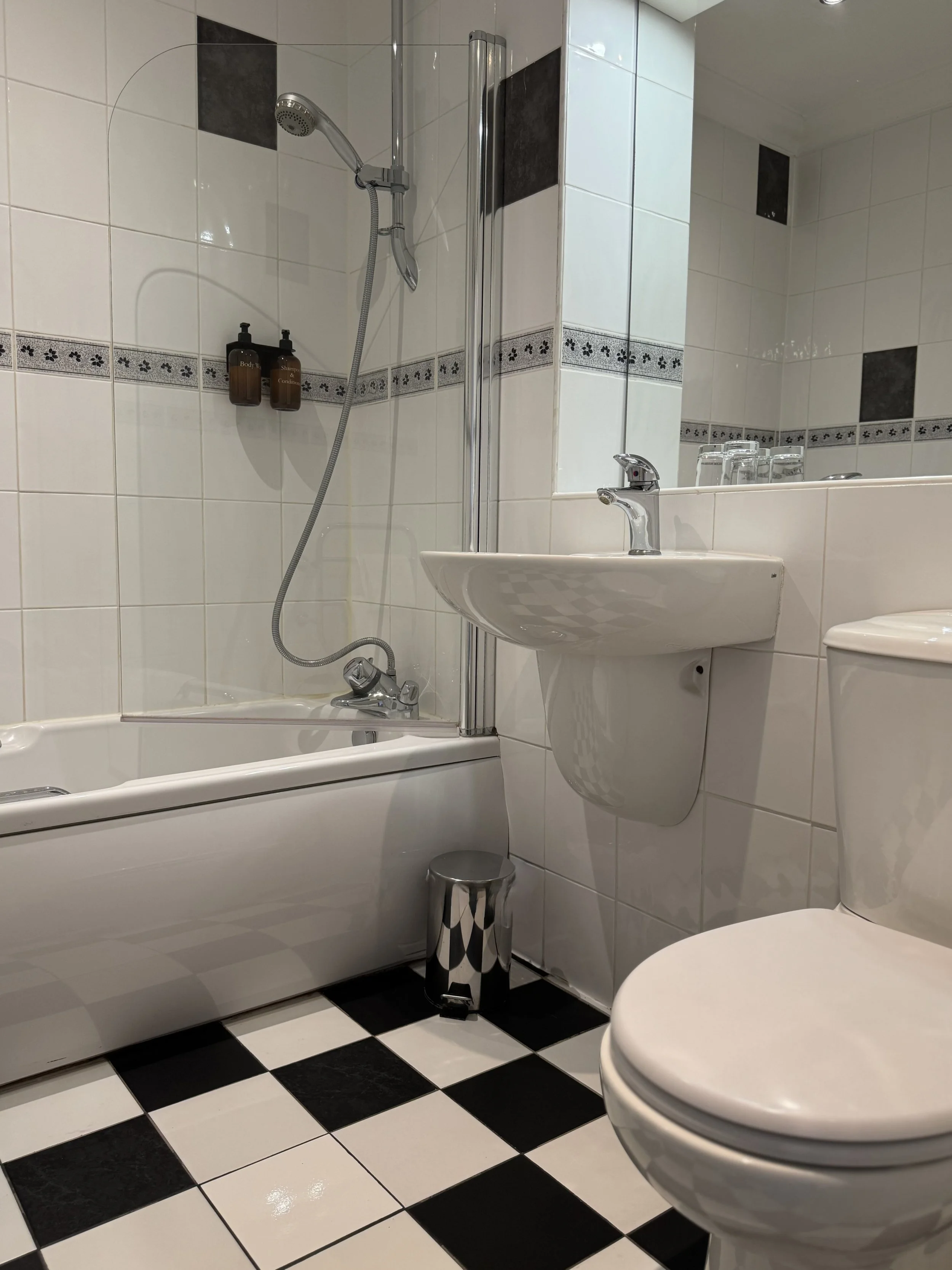 A small bathroom with a bathtub and shower combination, a sink with a mirror above, and a toilet. The floor has black and white checkered tiles, and there are two brown bottles on the shower wall.