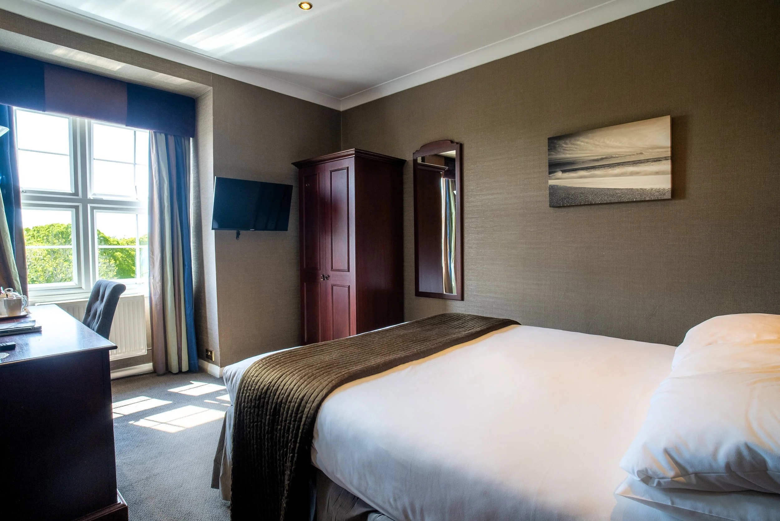 A hotel room with a bed, a window with curtains and a view of green trees outside, a wall-mounted TV, a wooden wardrobe, a mirror, and a framed black-and-white beach landscape picture.