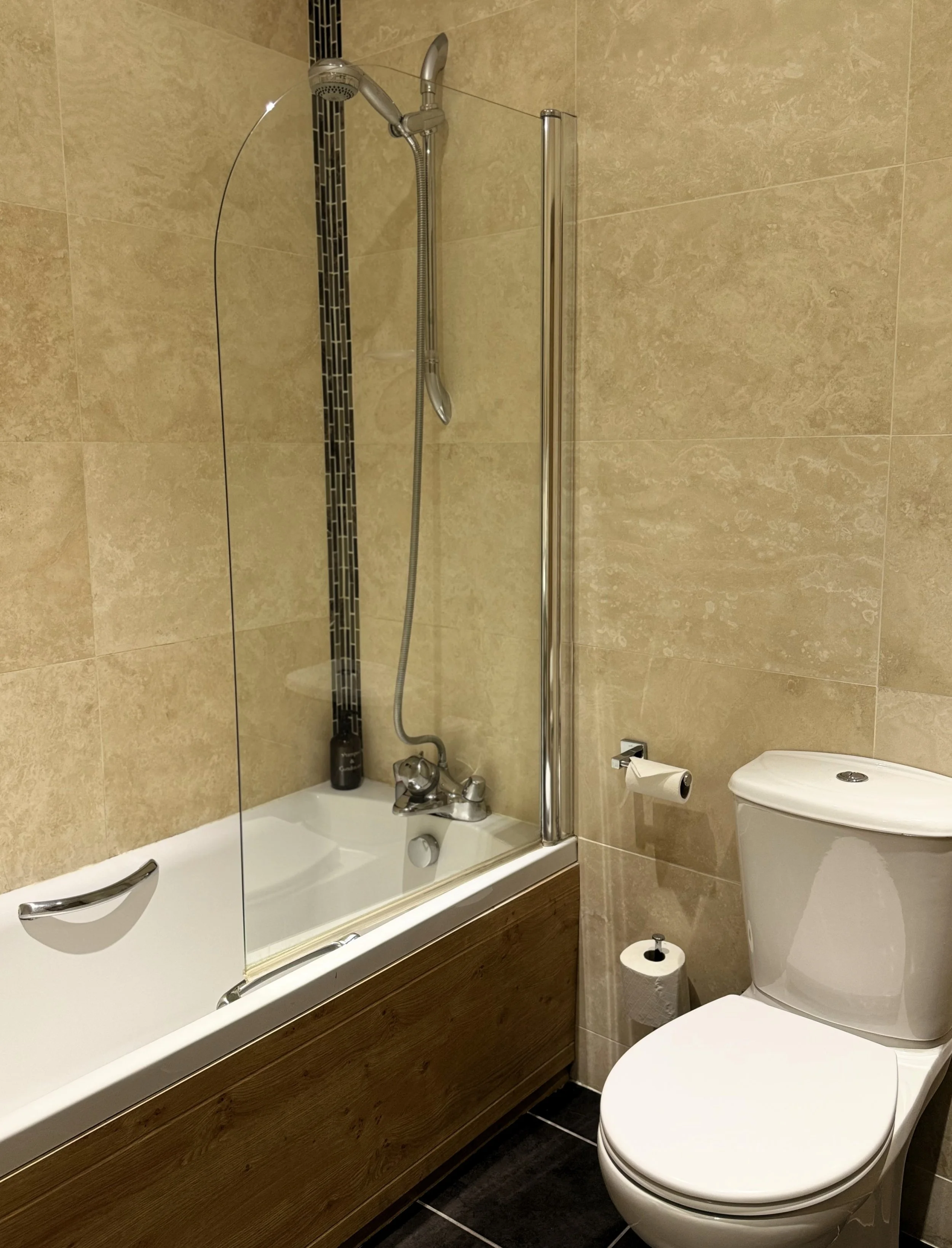 Bathroom with bathtub, shower, toilet, beige tiled walls, and black tiled accent.