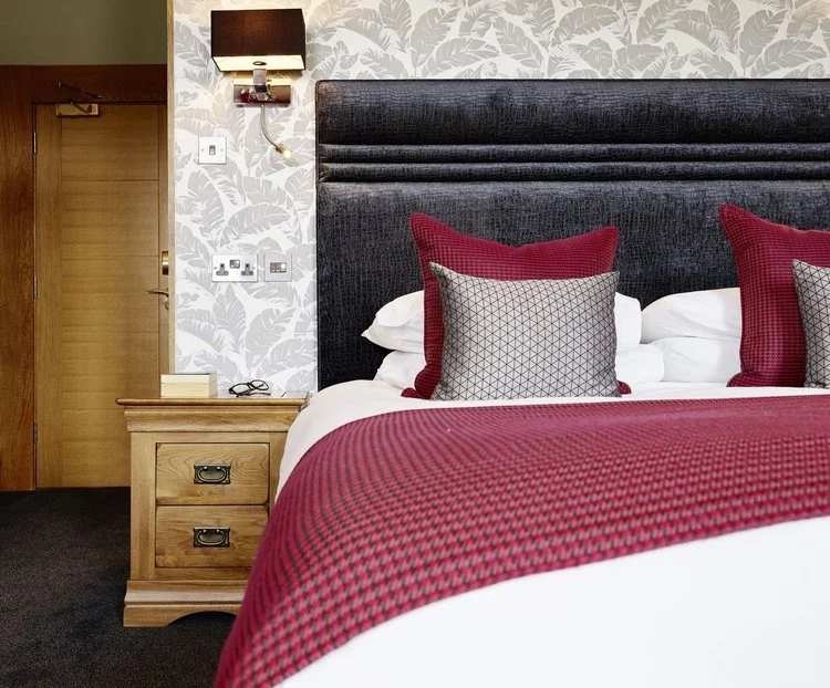 A hotel room with a bed, dark upholstered headboard, red and patterned throw pillows, and a red bedspread. A wooden nightstand with drawers is beside the bed, with a lamp, a box, and glasses on top. Wallpaper with a leaf pattern decorates the wall be