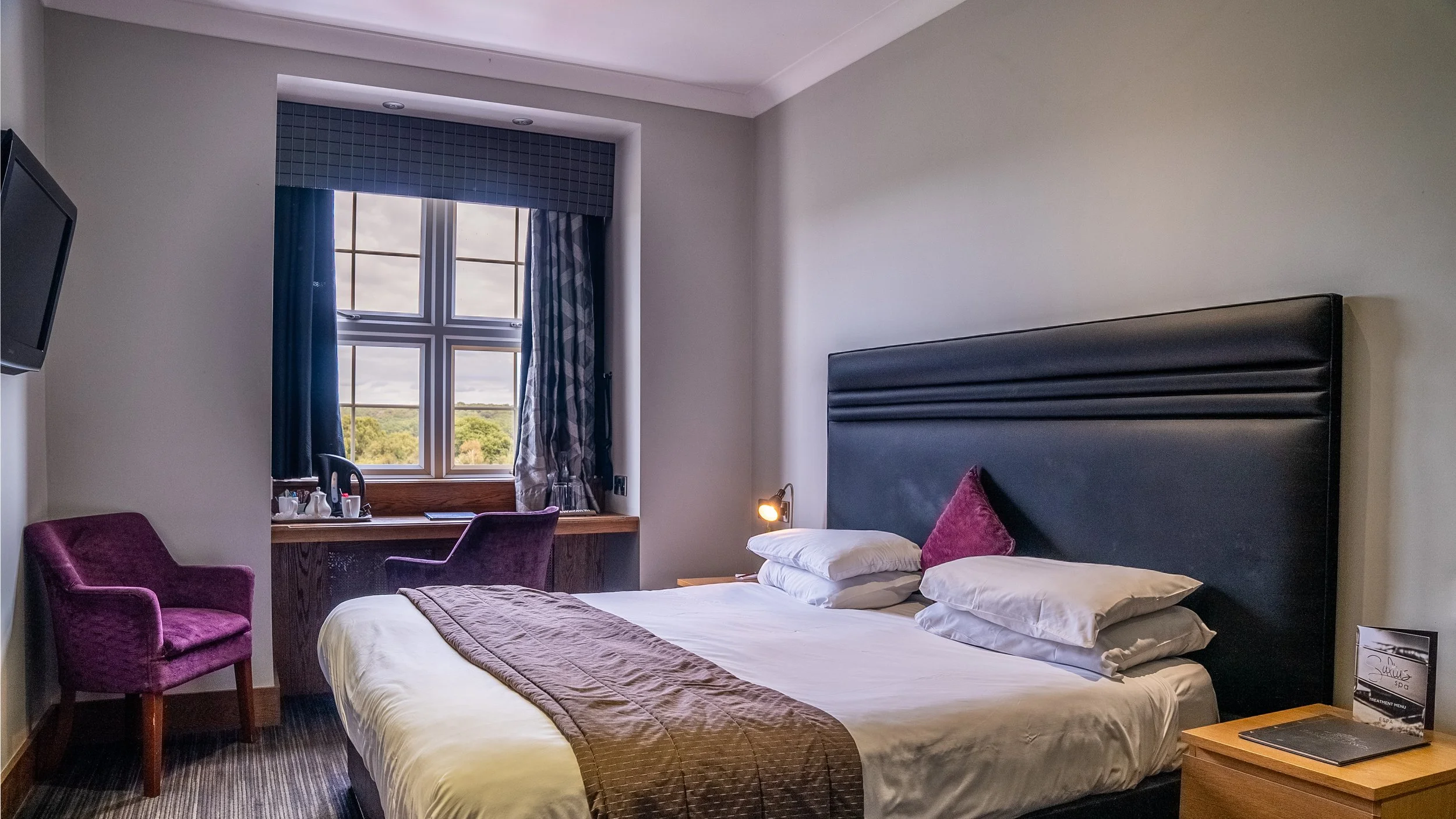 A hotel room with a large bed, purple armchair, window with curtains, and a desk with a chair inside.