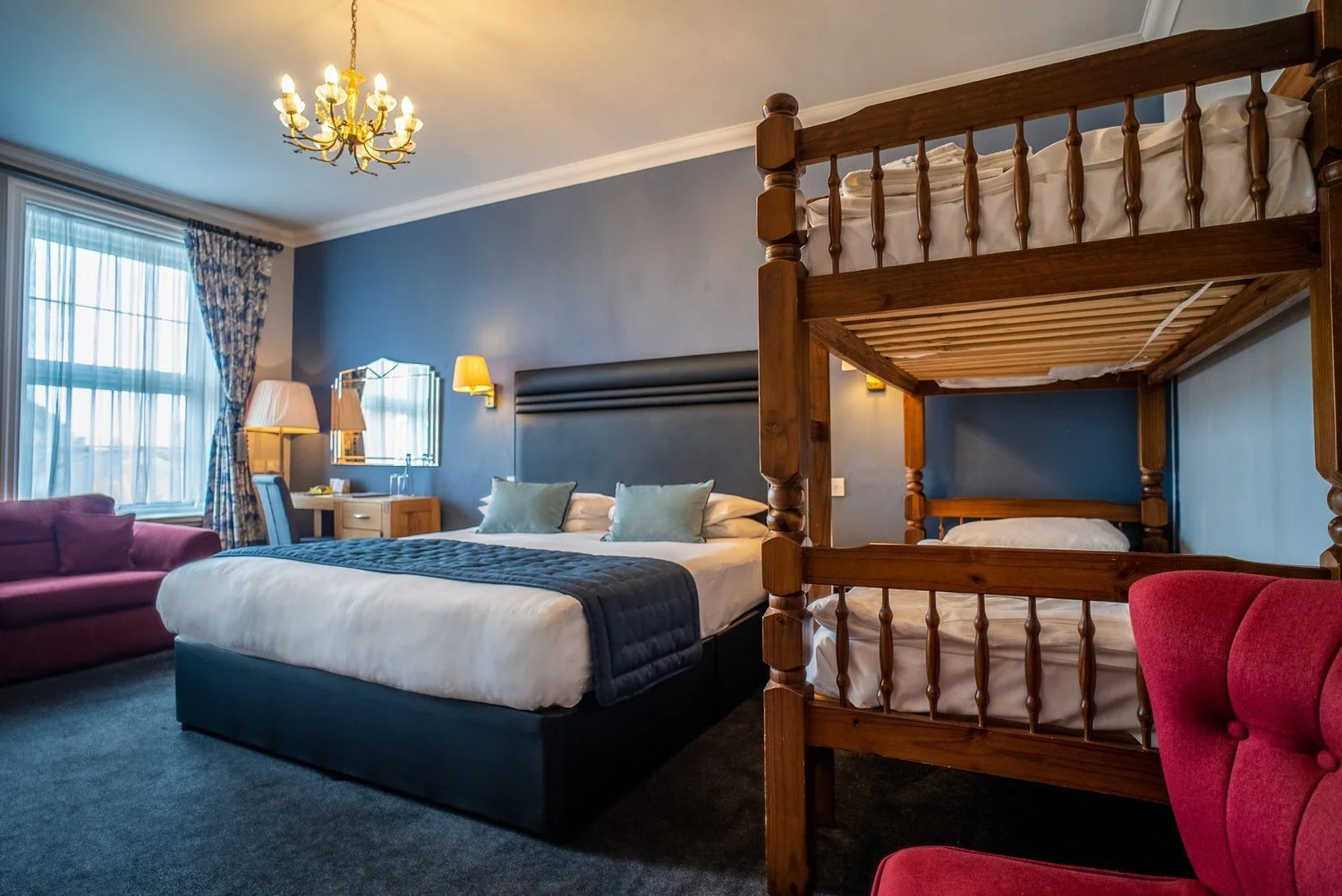 A hotel room with a large bed, a bunk bed, a pink armchair, a window with curtains, a desk with a chair, and a chandelier hanging from the ceiling.