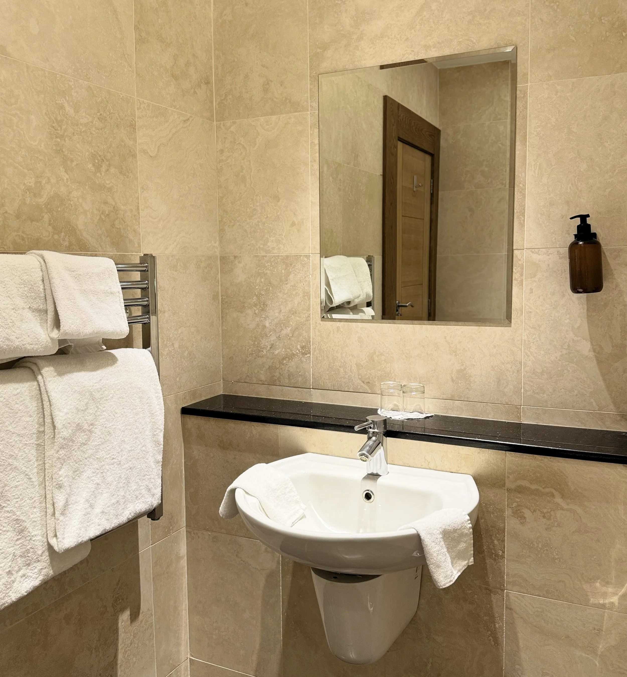 A bathroom with beige tiled walls, a wall-mounted white sink with a chrome faucet, a mirror above the sink, a brown soap dispenser on the wall, a towel rack with folded white towels, and two glasses on the black countertop.