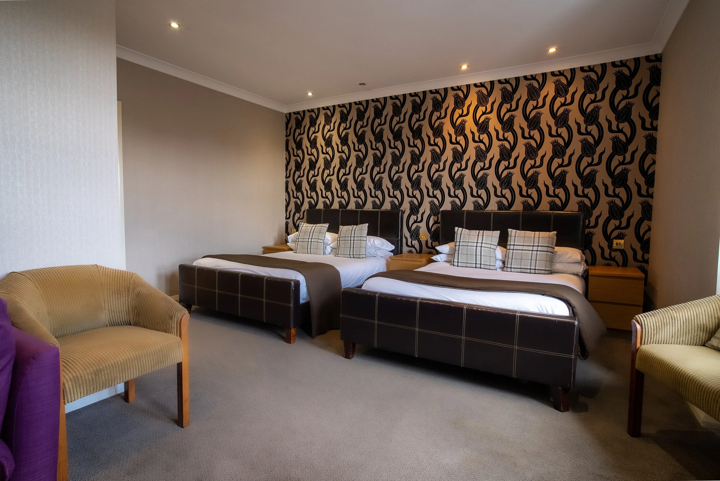 A hotel room with two double beds, black headboards, dark-patterned accent wall, beige armchair, and light-colored chair, with wooden nightstands and modern lighting.