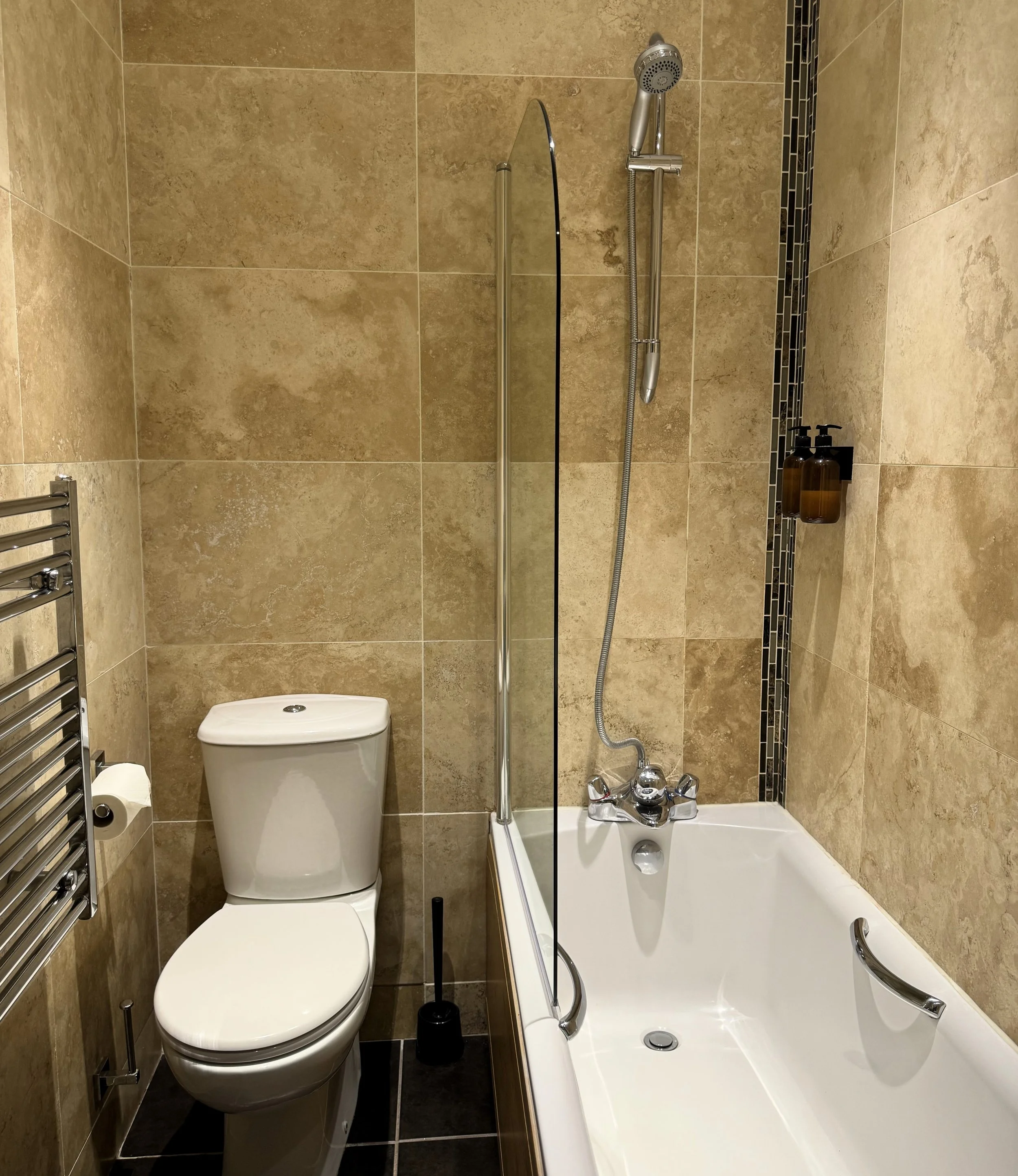 A small bathroom with beige tiled walls, featuring a toilet, a bathtub with a glass shower screen, a handheld showerhead, a towel radiator, a toilet brush, and a wall-mounted soap dispenser.