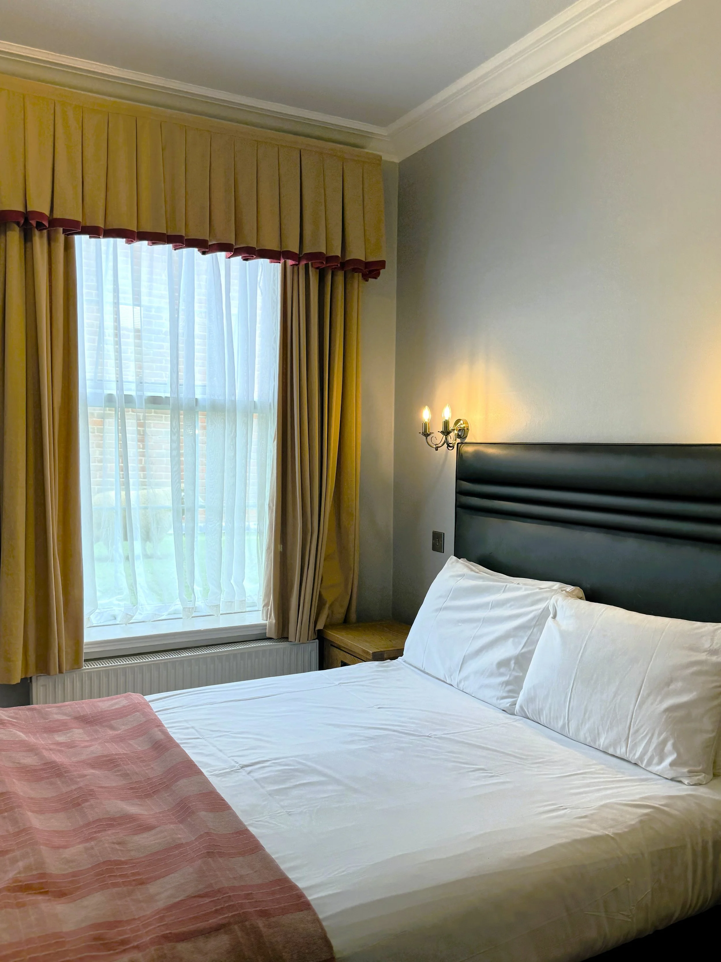 A neatly made bed with white sheets and pillows, a pink blanket at the foot, a window with beige curtains and sheer white drapes, a wooden nightstand, and a wall-mounted light fixture with two bulbs in a well-lit hotel room.
