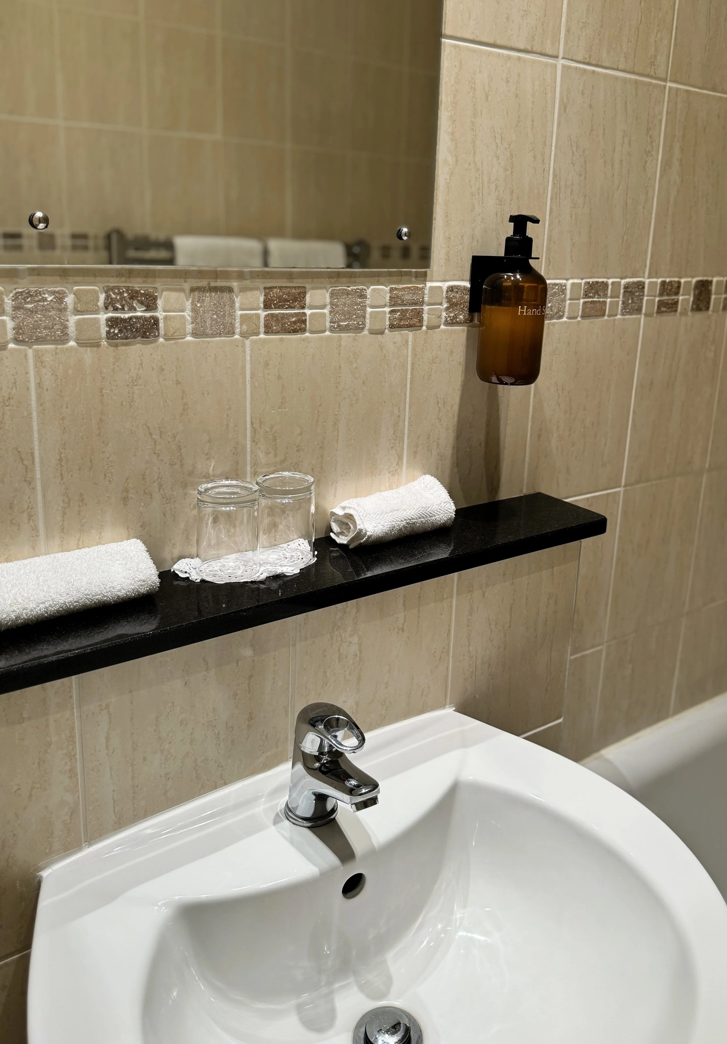 Bathroom sink with a silver faucet, a black shelf holding two rolled white towels and two upside-down glasses, a brown liquid soap dispenser, and a mirror above the sink.