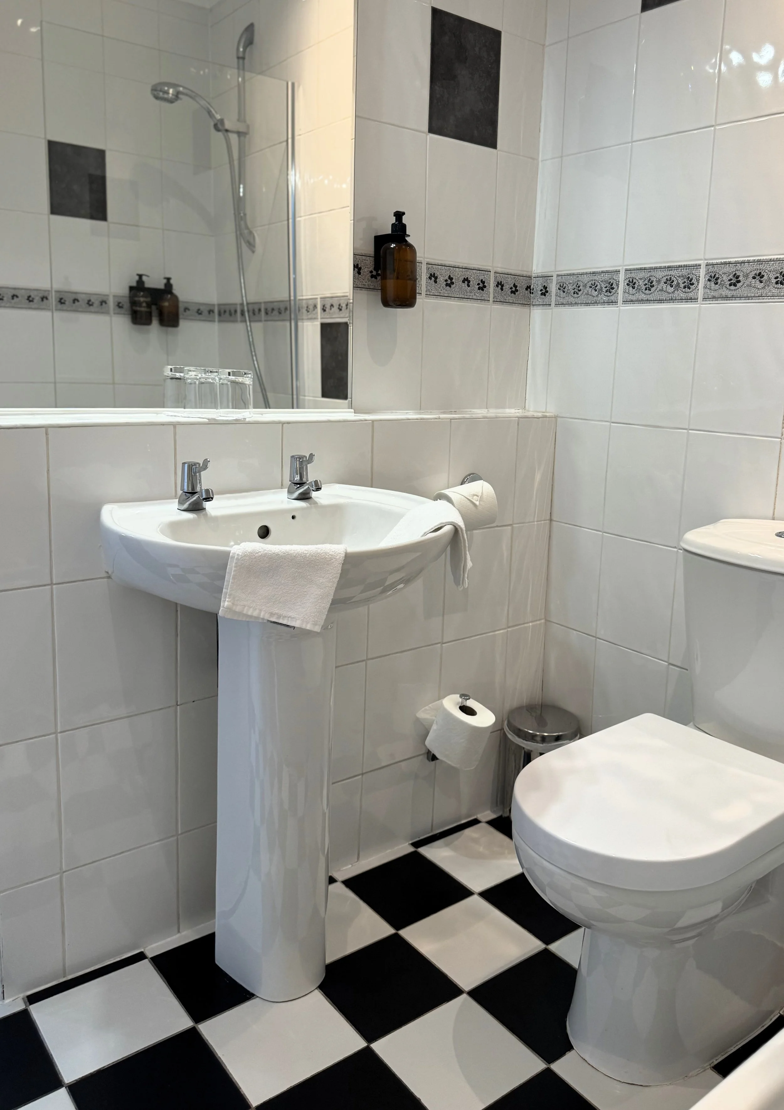 A small bathroom with white tiled walls, a pedestal sink with two faucets, a toilet, and a large mirror. There are black and white checkered floor tiles, a wall-mounted soap dispenser, a toilet paper holder, and a small trash can.