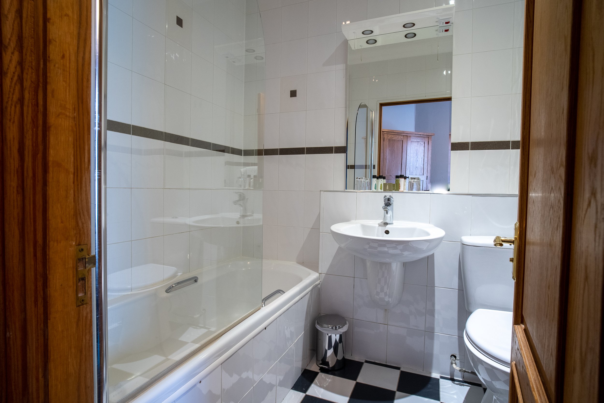 Small bathroom with a bathtub, sink, and toilet. White tiled walls with a horizontal band of dark tiles, checkered black and white floor, wood door, and a mirror above the sink.