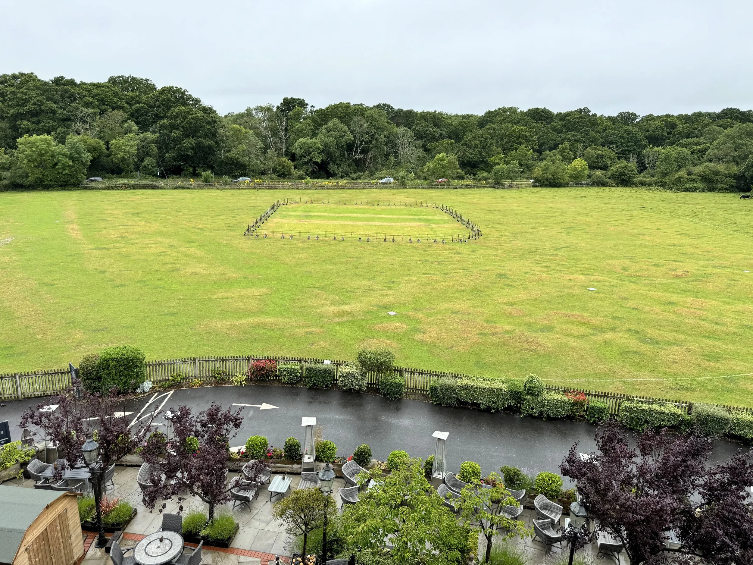A grassy field with a rectangular area marked by small stakes. In the foreground, there is an outdoor seating area with tables, chairs, trees, and patio heaters, separated by a wooden fence, with a parking lot behind the fence.