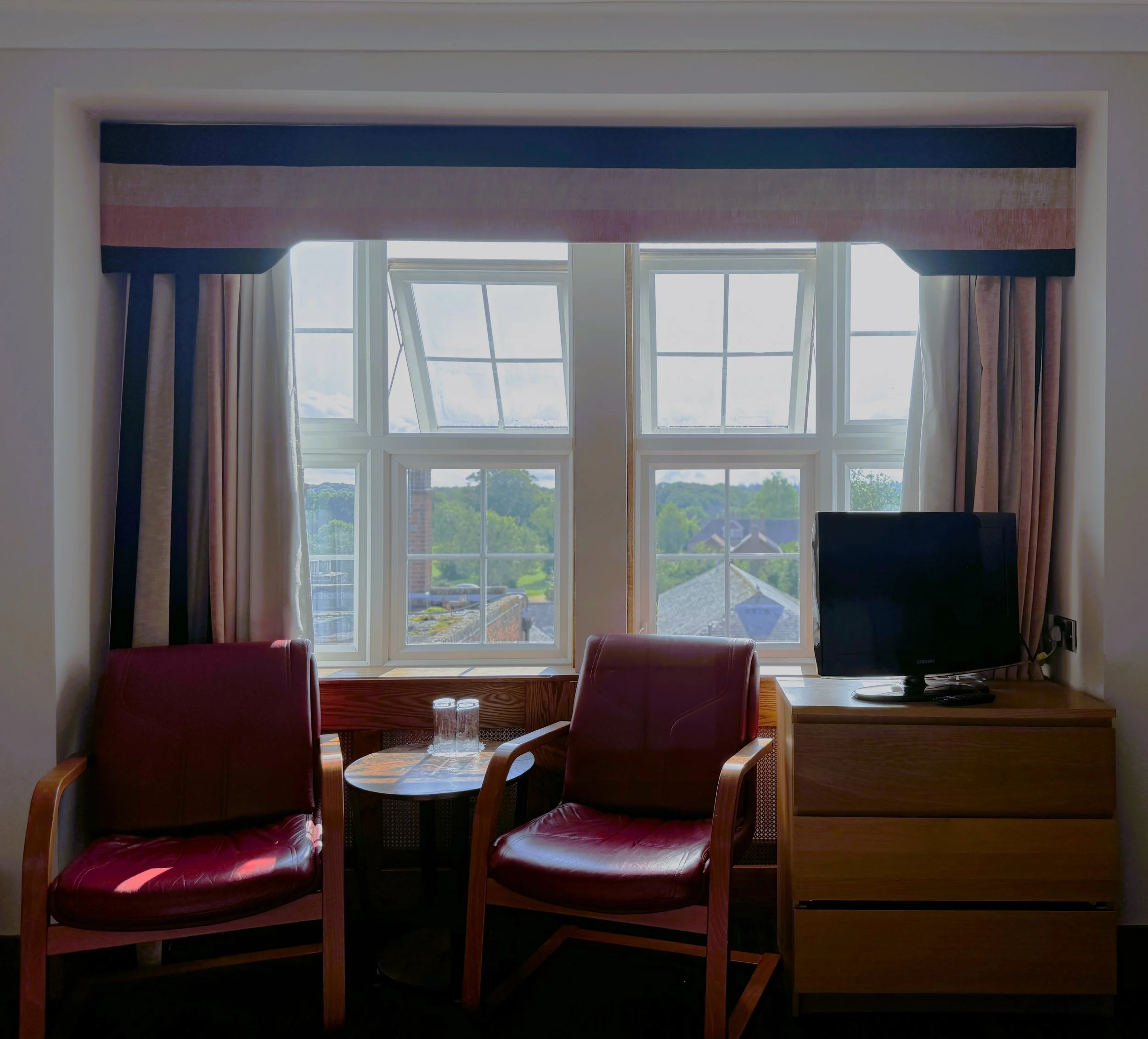 Hotel room with two red leather chairs, a small round wooden table with two glasses, a window with curtains, and a television on a wooden dresser.