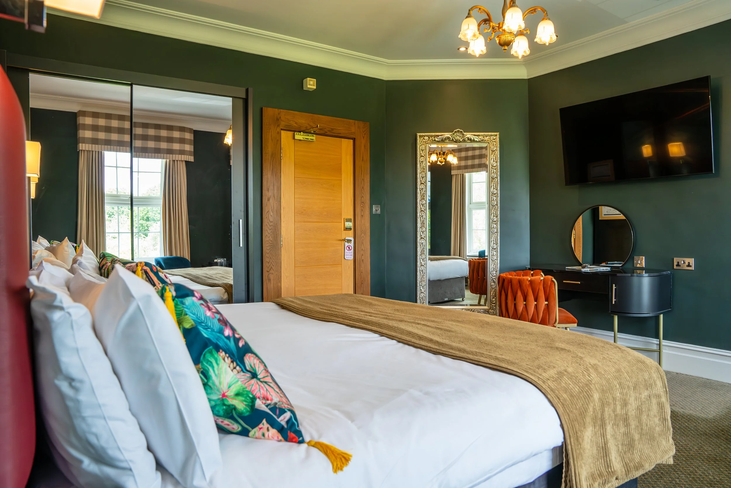 A cozy hotel bedroom with a bed decorated with colorful pillows and a gold-colored bedspread, dark green walls, a large mirror, a mounted flat-screen TV, and a vintage-style desk with an orange tufted chair.