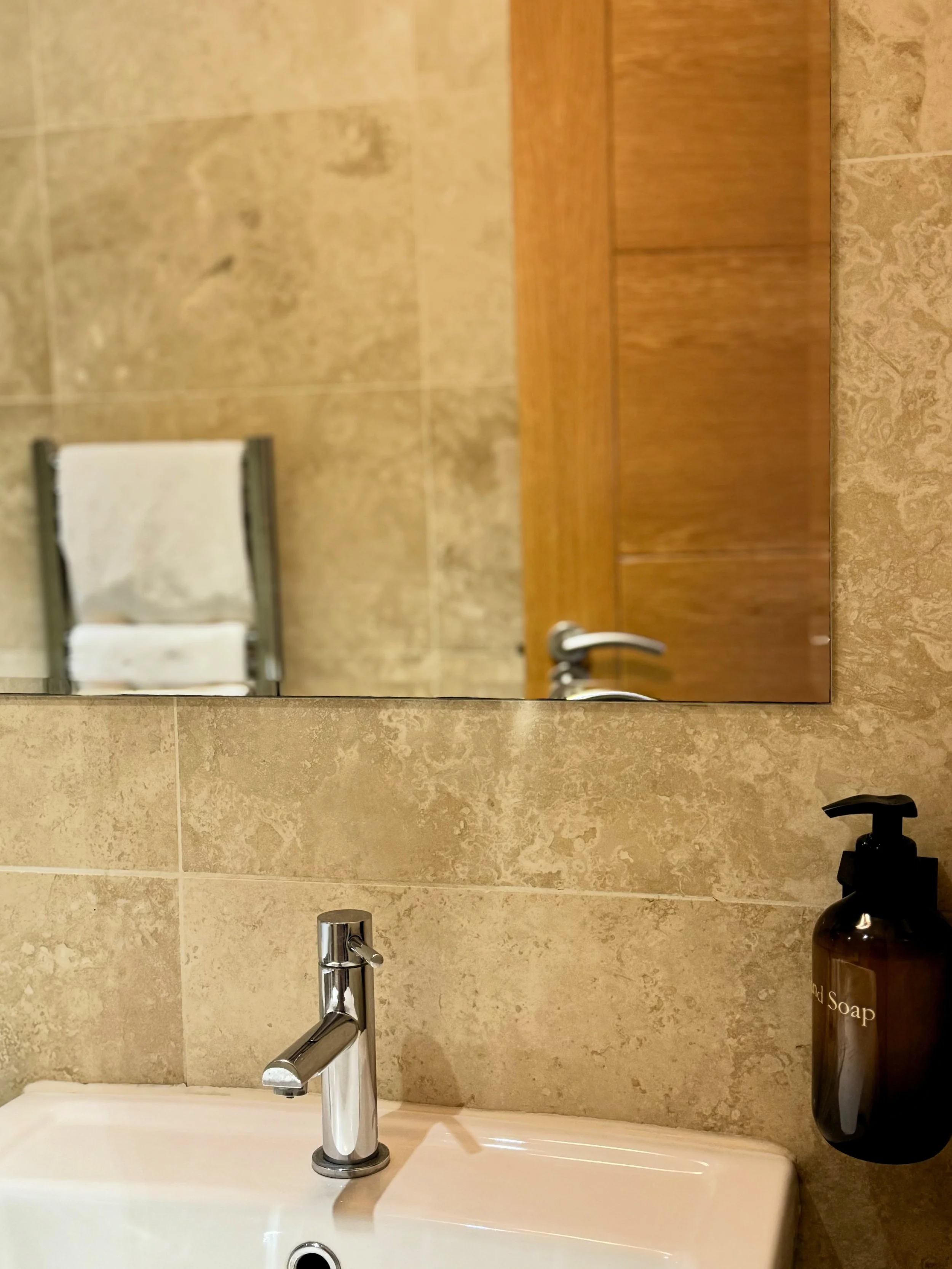 Bathroom sink with chrome faucet, soap dispenser labeled 'Hand Soap,' tiled wall, mirror reflecting towels on a rack, wooden cabinet, and door handle visible.