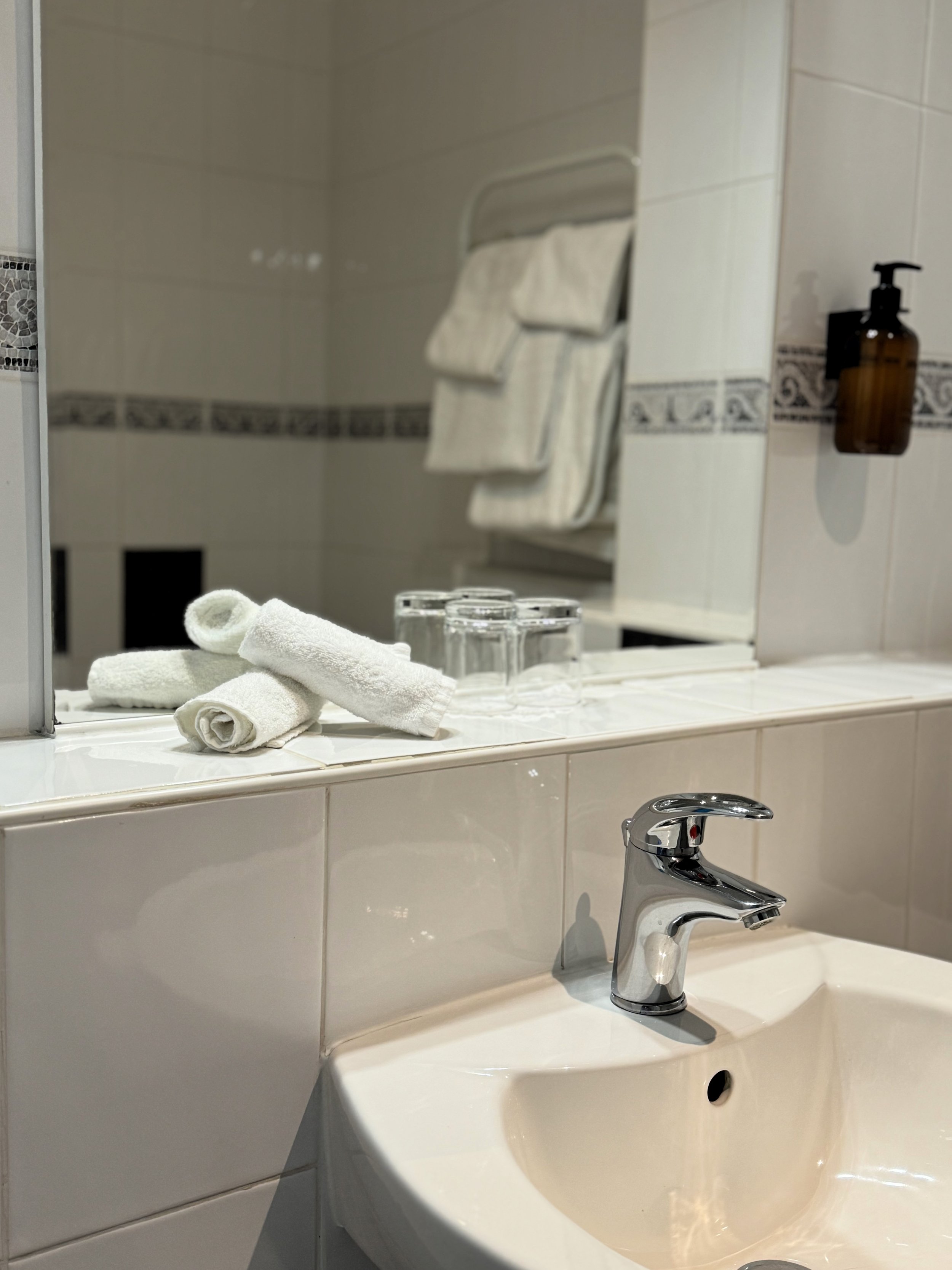 Bathroom countertop with folded white towels, four upside-down glasses, sink, and chrome faucet, with mirror reflecting towels and wall-mounted soap dispenser.