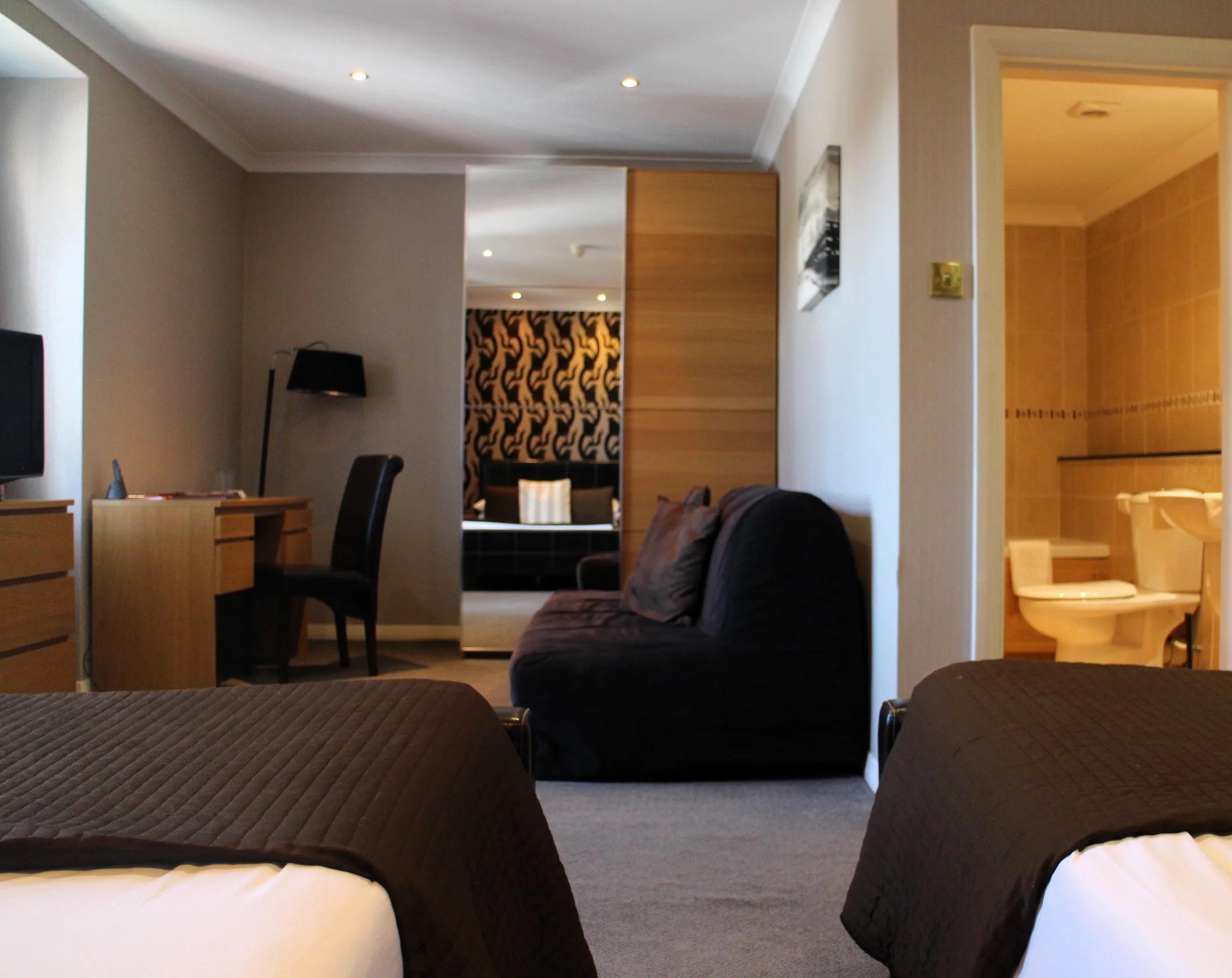 Hotel room with two beds, a black sofa, a desk with a chair, a black floor lamp, a television, and a view into the bathroom.