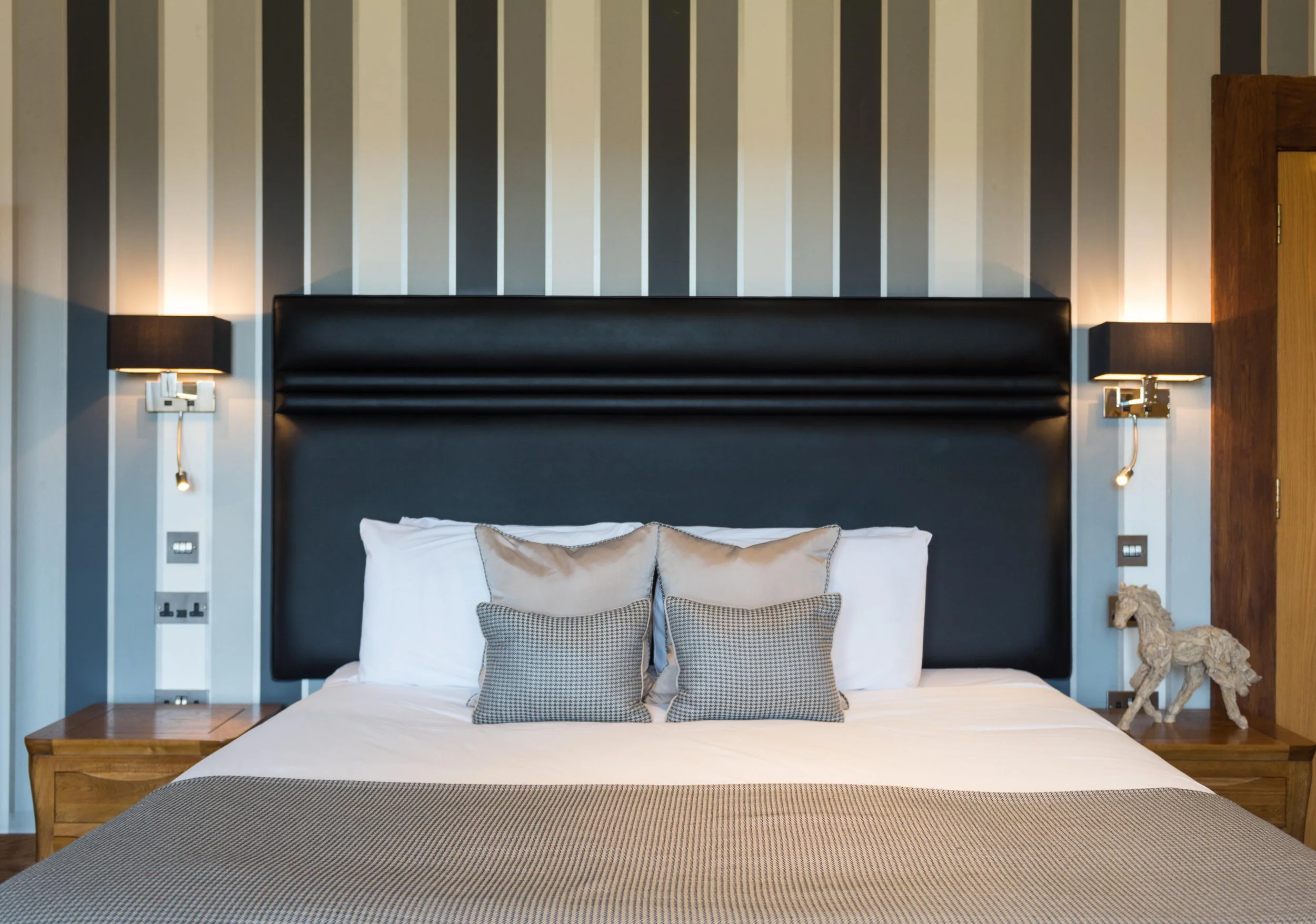 A neatly made bed in a modern hotel room with a dark headboard, striped wall behind, and paired with bedside tables and wall-mounted lamps. Four decorative pillows are arranged on white bedding, with a beige runner at the foot of the bed.