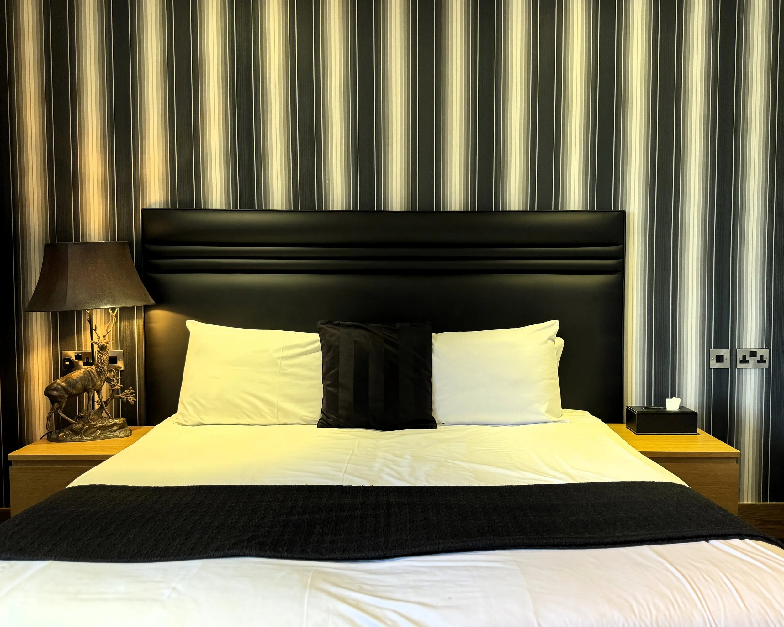 A neatly made bed with white sheets, two white pillows, a black headboard, and a black decorative pillow. On each side of the bed are wooden nightstands; the left has a lamp and a decorative deer sculpture, and the right has a tissue box. The wall be