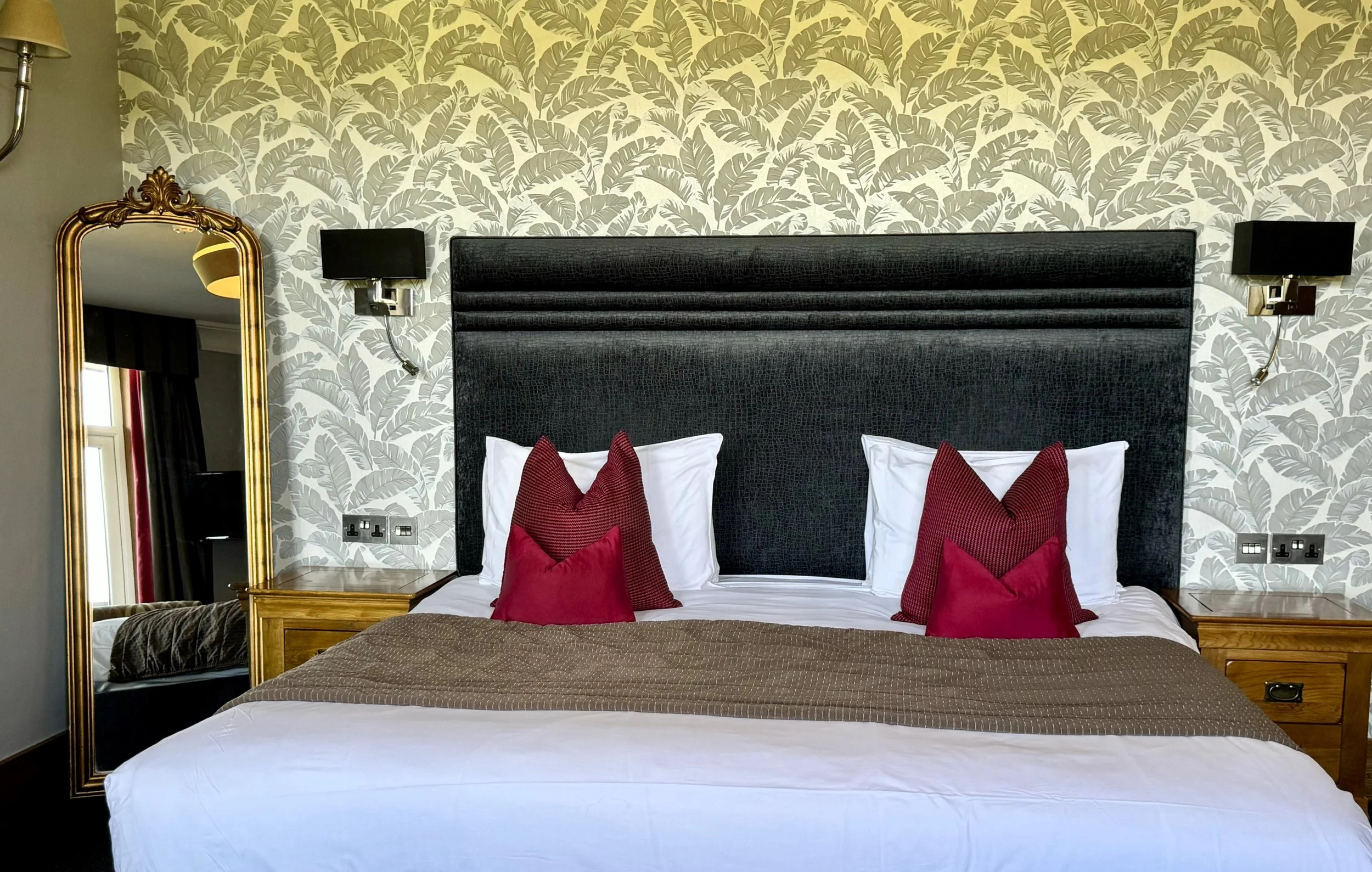 Hotel room bed with black upholstered headboard, white bedding, red and brown accent pillows, wooden bedside tables, wall-mounted black lamps, and a decorative leafy wallpaper.