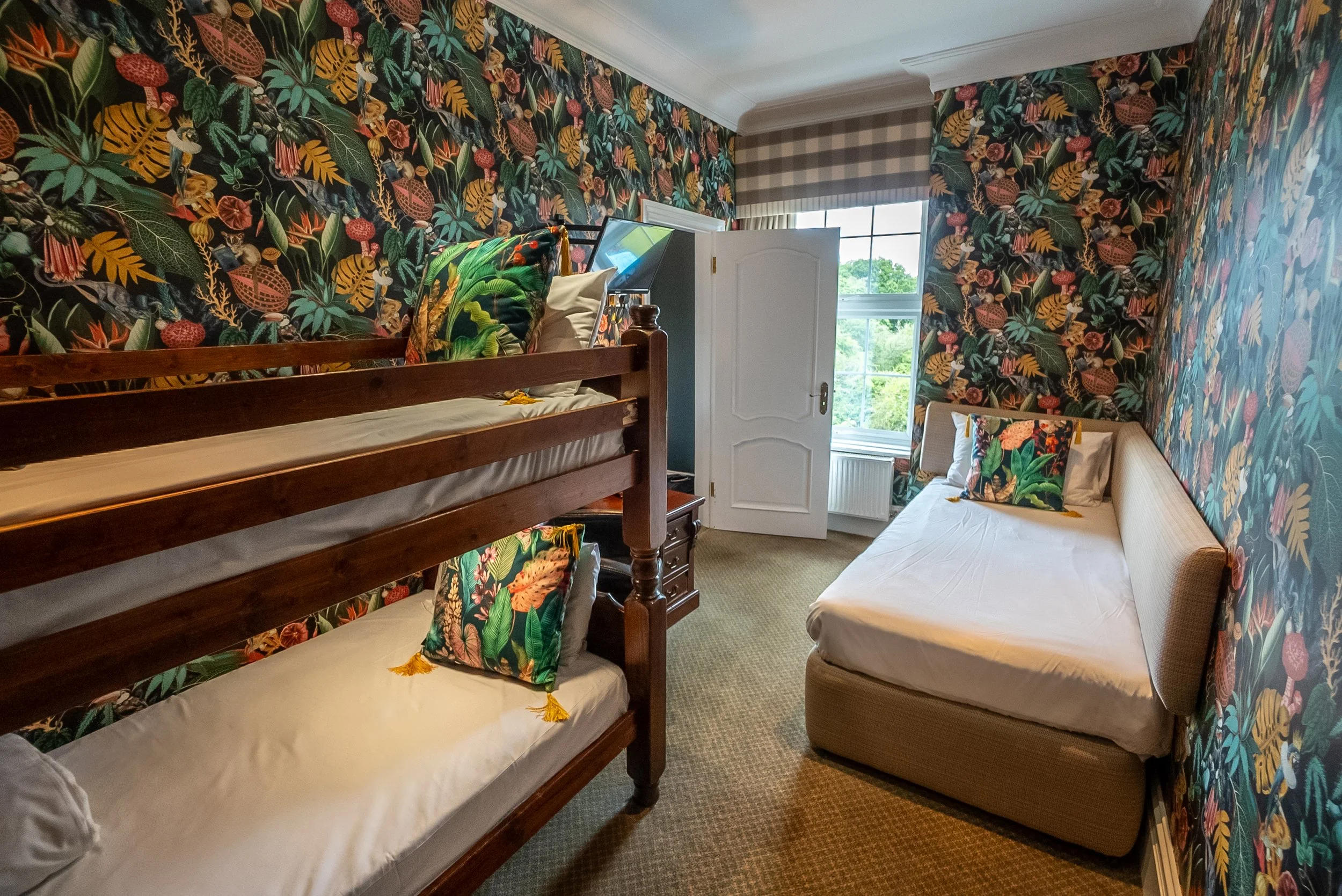 Twin bunk beds and a daybed in a room with vibrant tropical-plant-themed wallpaper, a window with blinds, and assorted pillows.