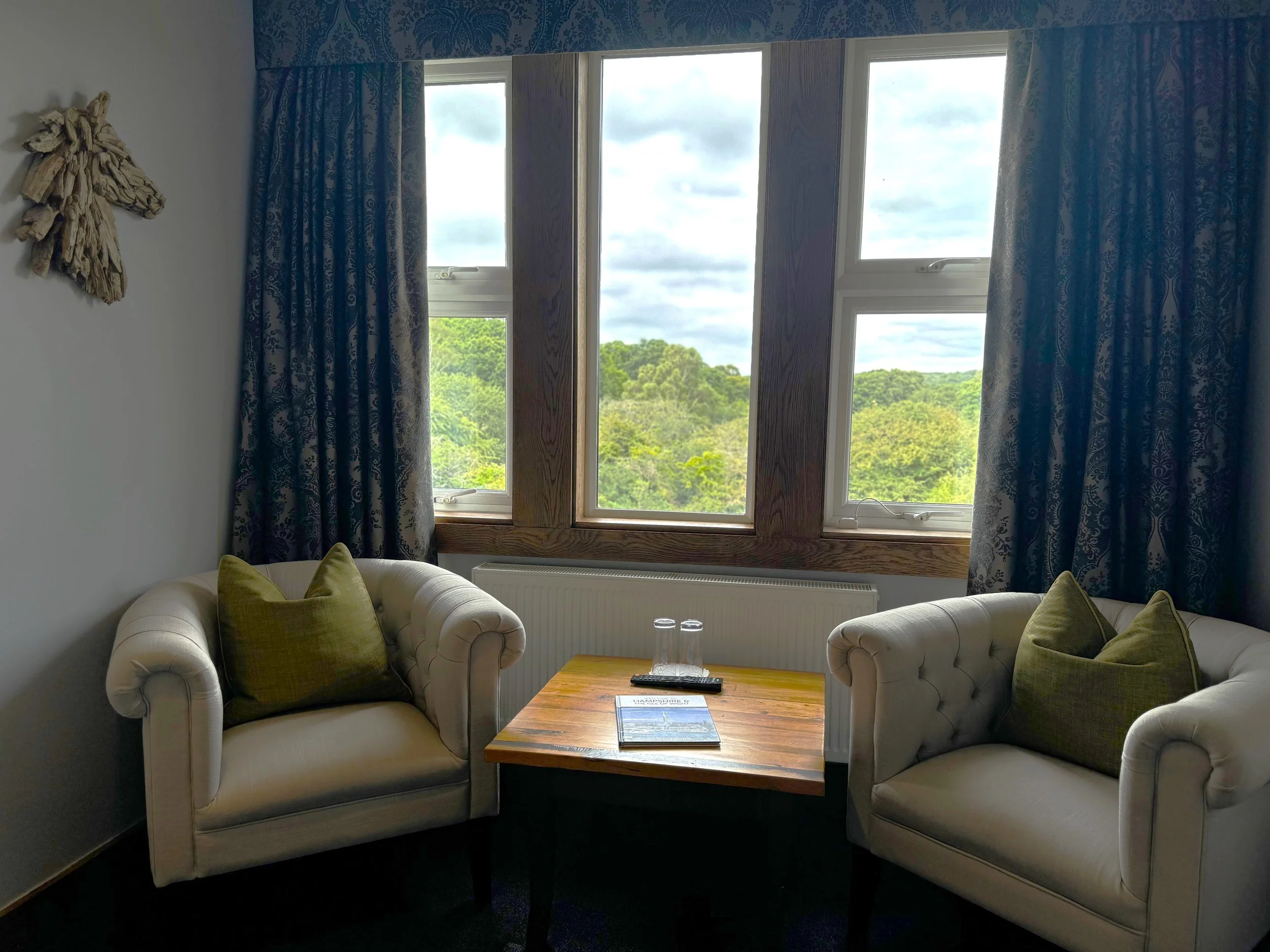 A cozy living room corner with two white tufted armchairs with green pillows, a wooden side table with glasses and a magazine, large multi-pane windows with dark patterned curtains, and a view of green trees outside.