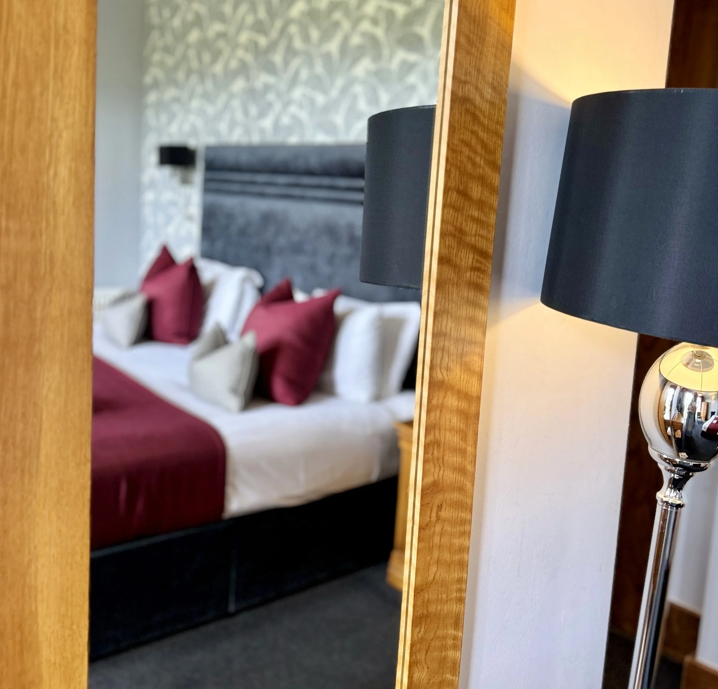 Close-up of a modern lamp with a black shade, reflected in a mirror, with a bedroom scene featuring a bed with white linens, maroon and white pillows, and a decorative wall in the background.