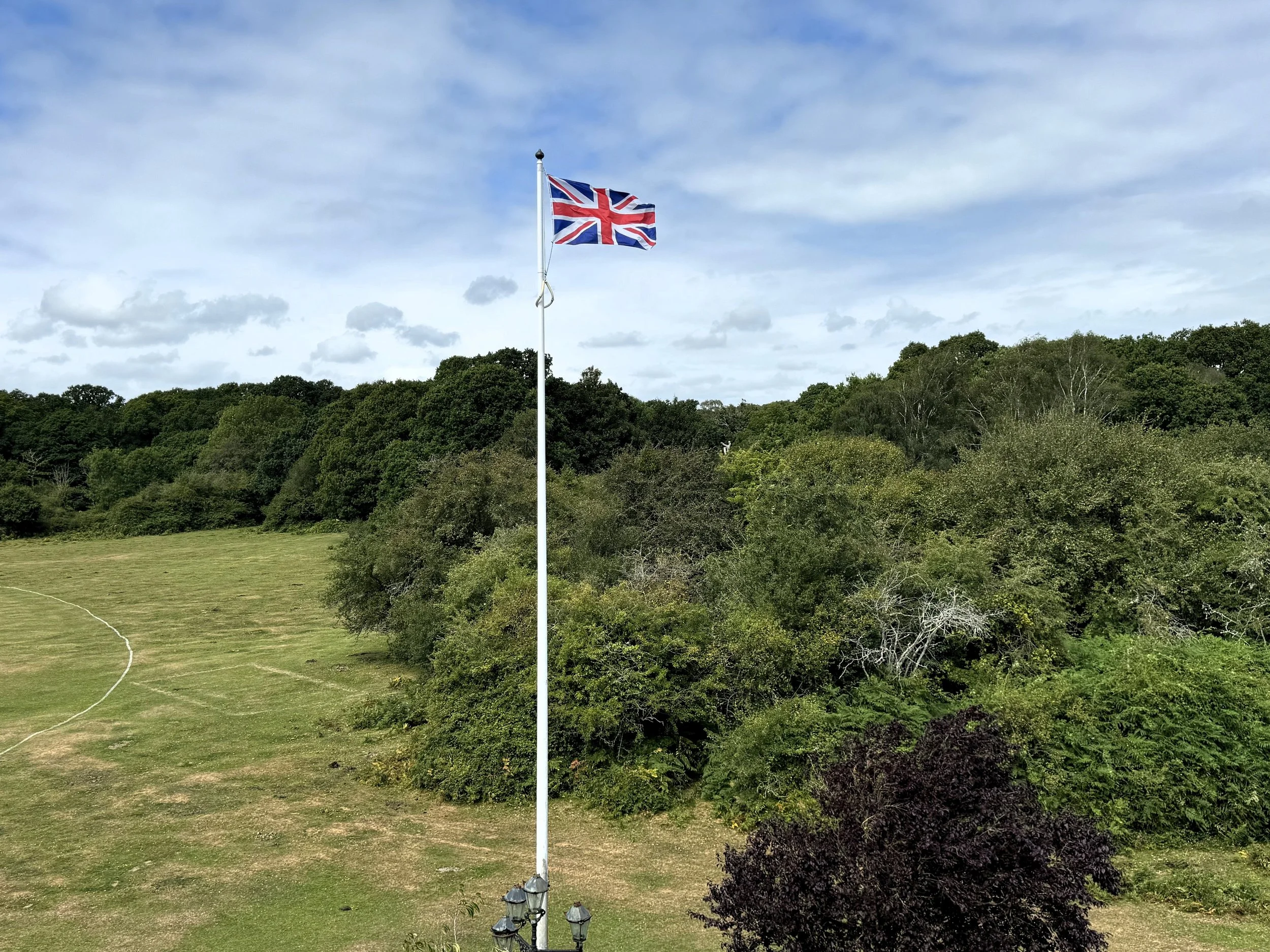 British flag flying on a tall flagpole in a park with trees and grass, under a partly cloudy sky.