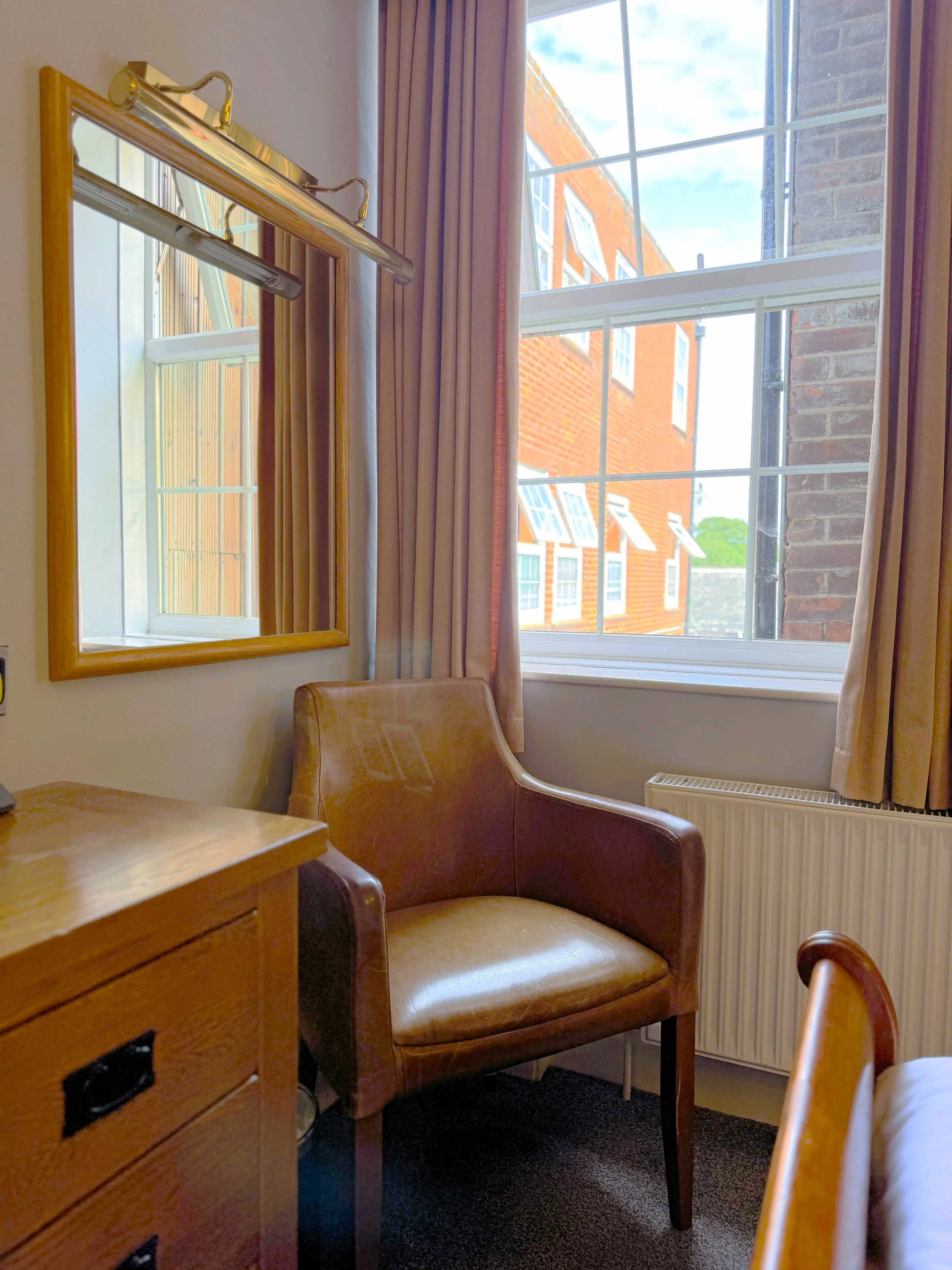 Interior of a room with a brown leather armchair near a window, with beige curtains, a wooden desk, a mirror with a light fixture, and a view of red-brick buildings outside.