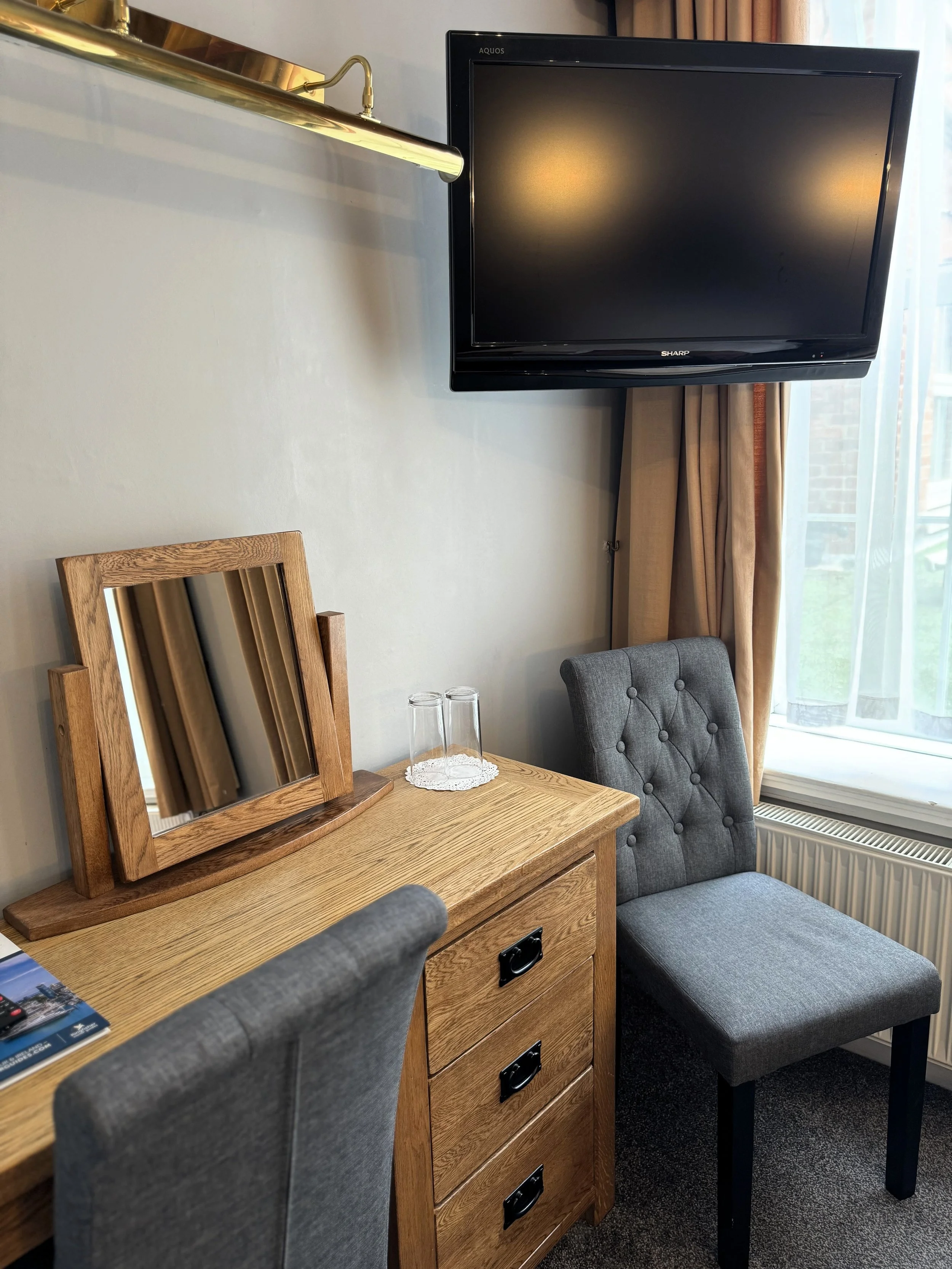 A hotel room corner with a wooden desk, a gray upholstered chair, a small gray tufted stool, a wall-mounted flat-screen TV, and a window with curtains.