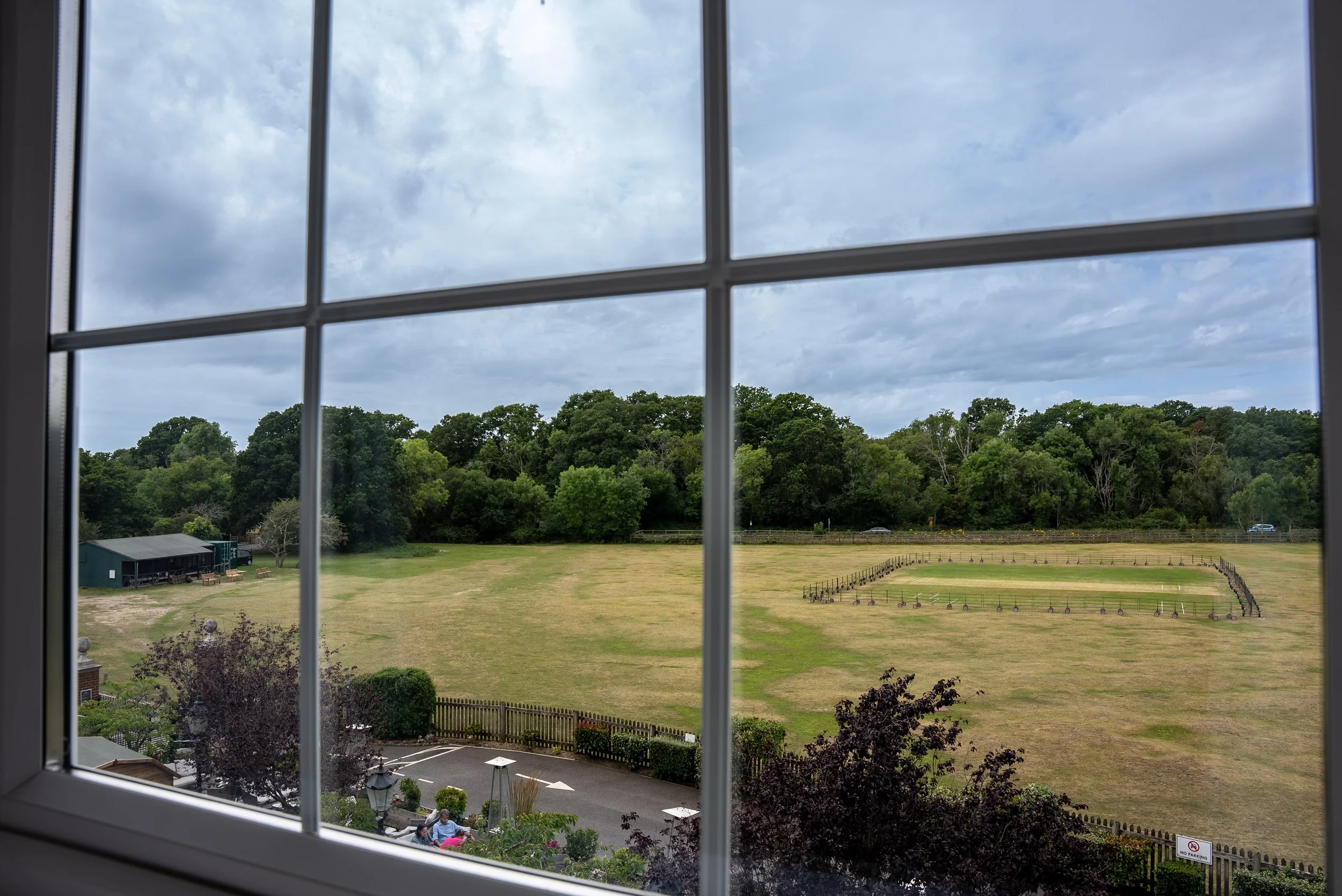 View of a large grassy field with a fenced-in area, cars parked near the fence, and a background of trees and cloudy sky, seen through a window with 8 panes.
