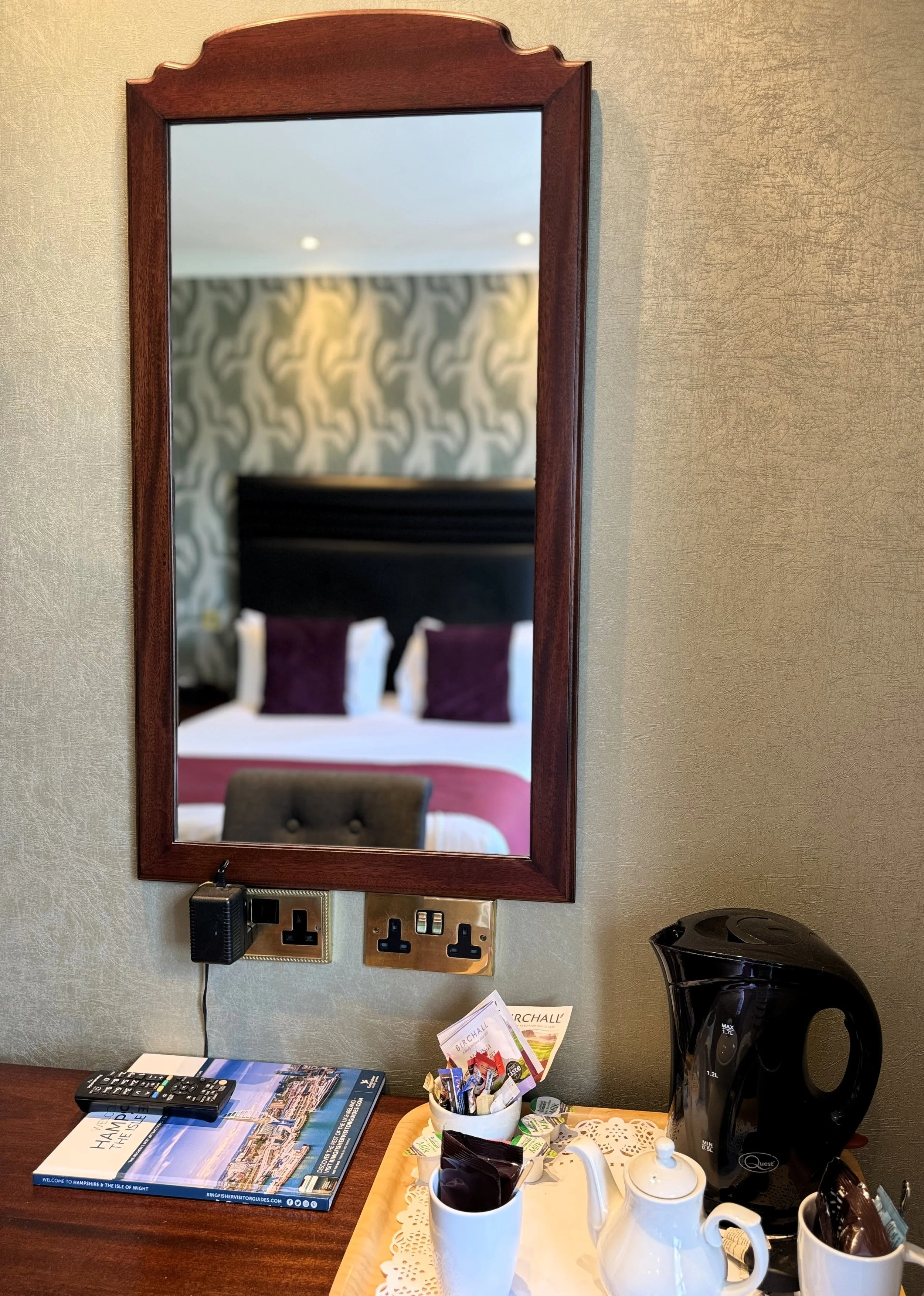 A hotel room with a wooden framed mirror reflecting a bed with purple pillows and a patterned wall. On the desk below the mirror, there is a remote, a travel brochure, a black adaptor, a tray with a black electric kettle, a white teapot, cups and sau