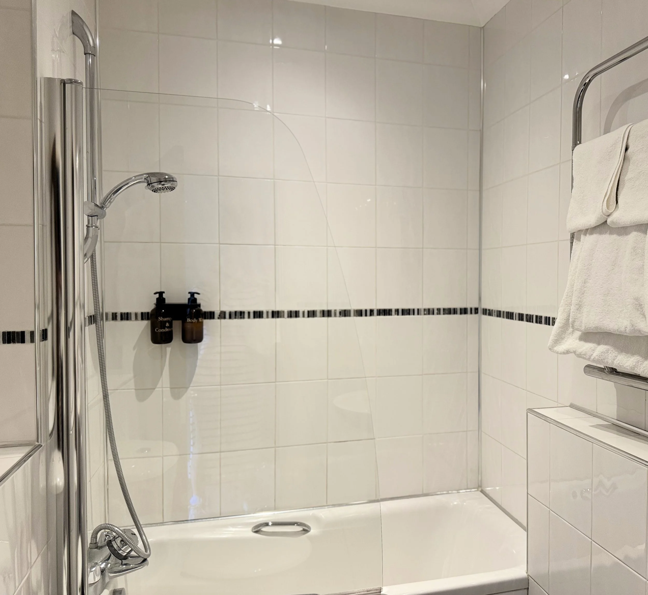 Empty white tiled shower with black accent tiles, shower head, two soap dispensers, and a towel hanging on a metal rack.