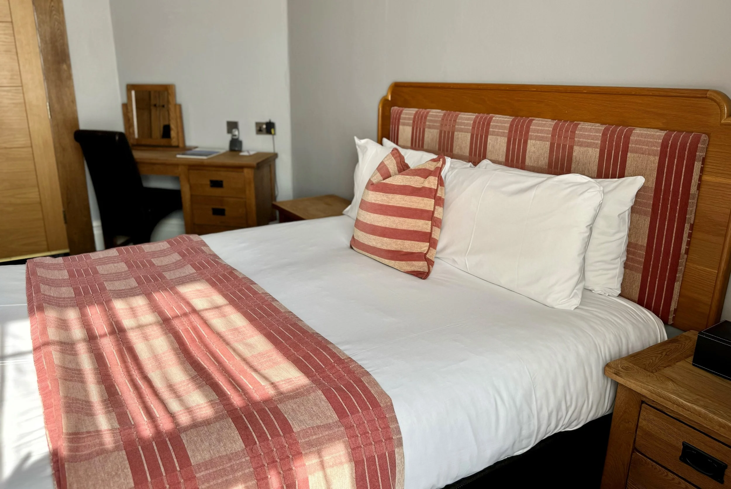 A neatly made bed with white linens, a striped pillow and bed runner, wooden headboard, next to a wooden nightstand, and a desk with a chair in the corner of the room.