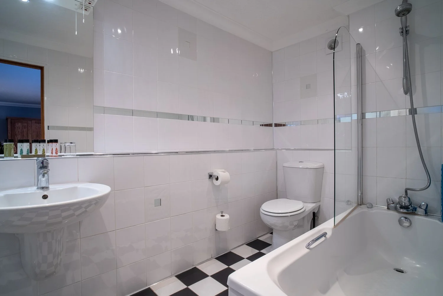 A clean bathroom with white tiled walls, a mirror, a toilet, a sink, and a bathtub with a shower, and black and white checkered floor tiles.