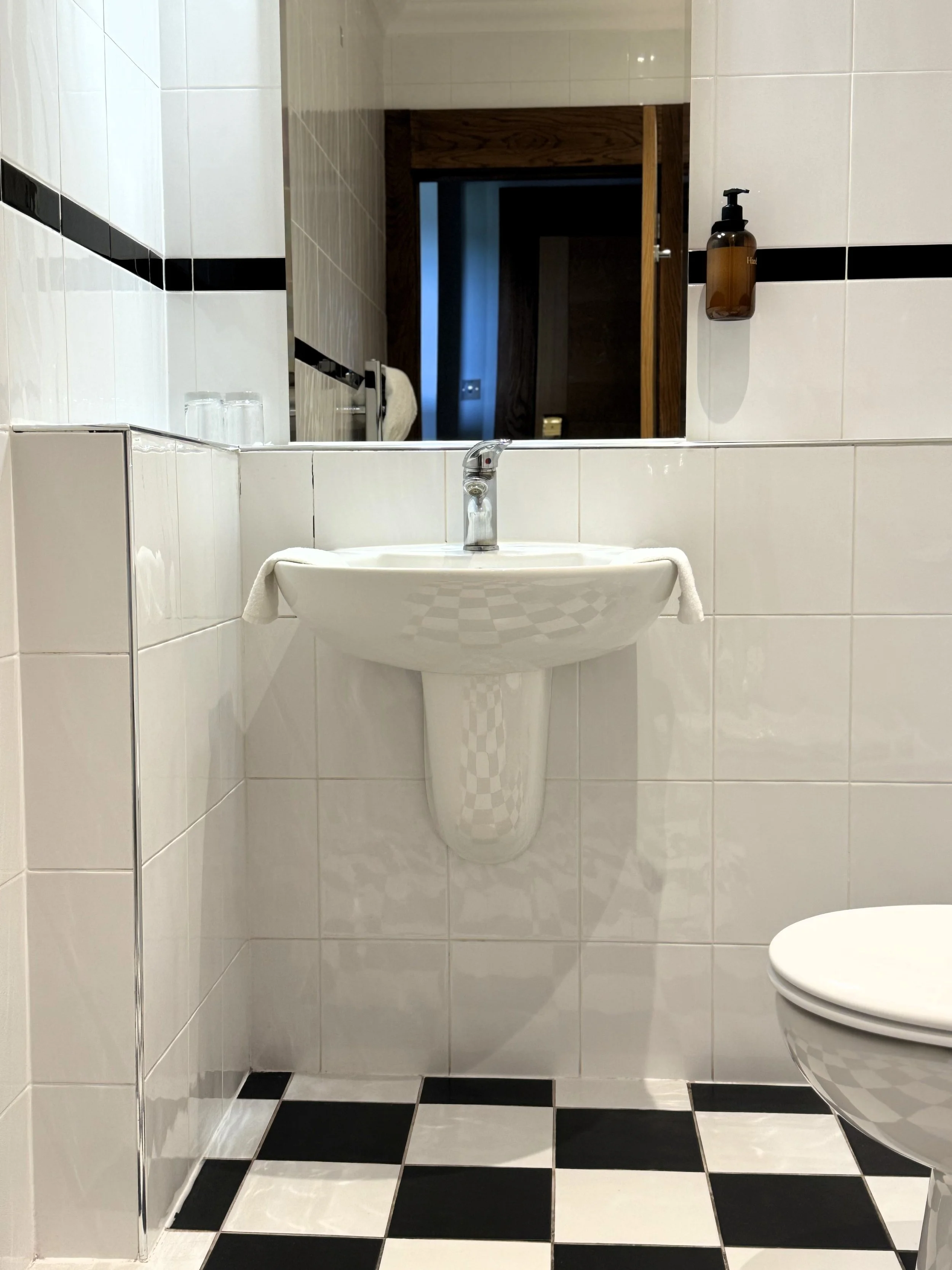 White bathroom with a sink, mirror, and toilet, black and white checkered floor tiles, and a dark brown soap dispenser on the wall.
