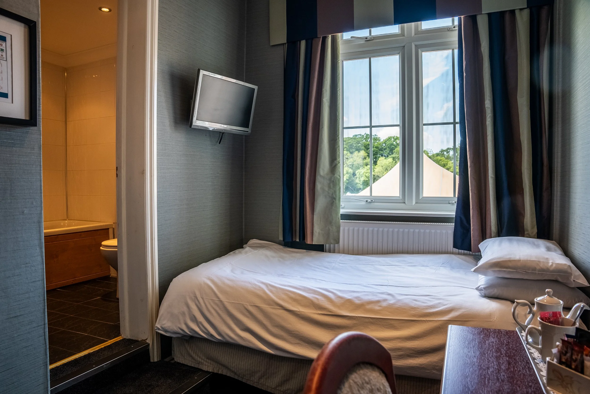 A hotel room with a bed next to a window, a mounted TV, and a view of trees outside. A bathroom with a toilet and bathtub is visible through an open door.